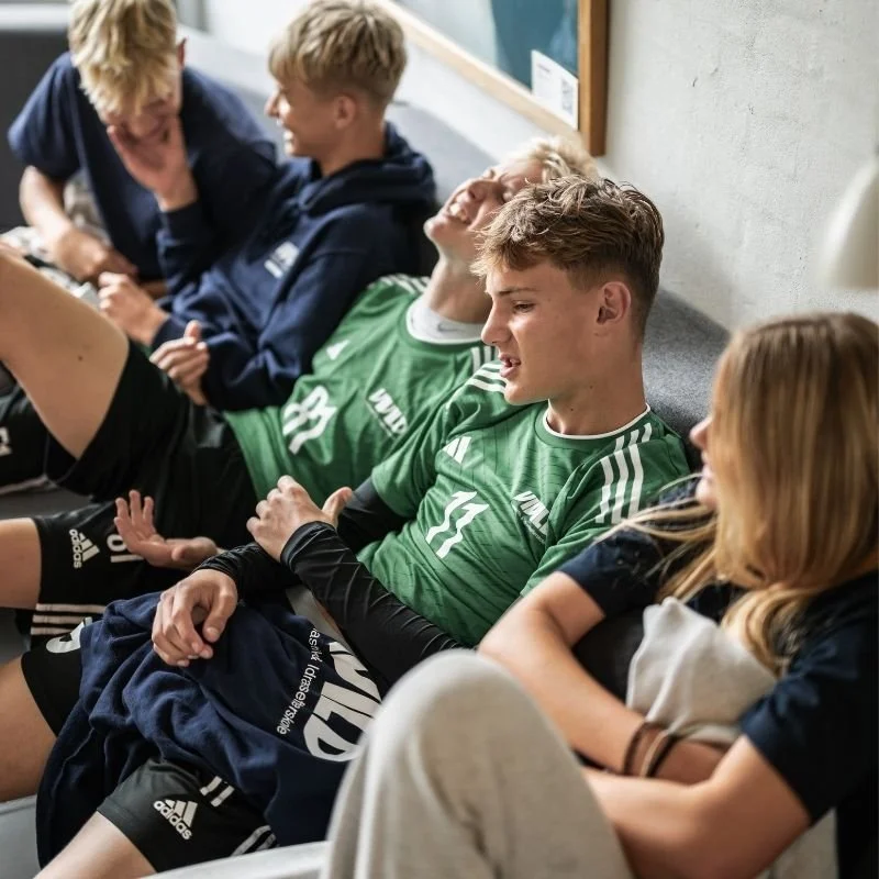 Fodboldspillere sidder på en bænk, nogle af dem griner og har afslappede positurer.