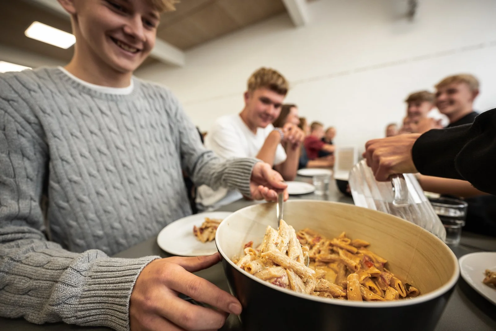 Smilende personer serverer pasta til en gruppe mennesker ved et langbord i en restaurant eller kantine.