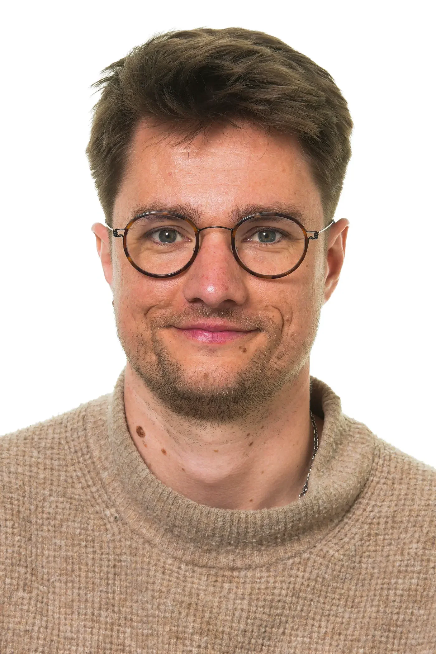 Et tæt portræt af en smilende mand iført runde briller og en blå trøje, med tydelige fregner og mærker på hagen.
