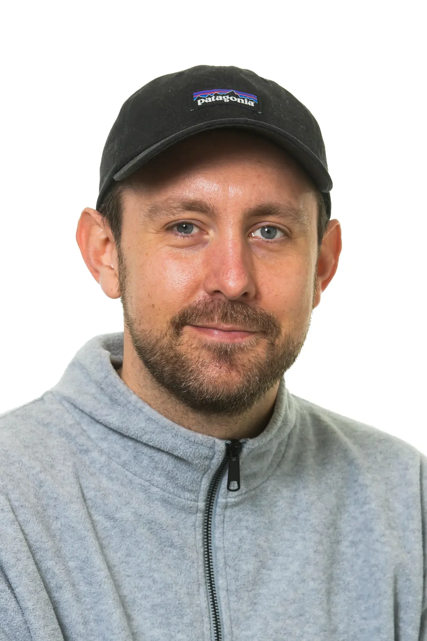 Menneske med blå øjne, lyser hud, kort brunt hår, lyst skæg, iført grå hoodie og sort baseballcap med teksten 'Jas Dou'.