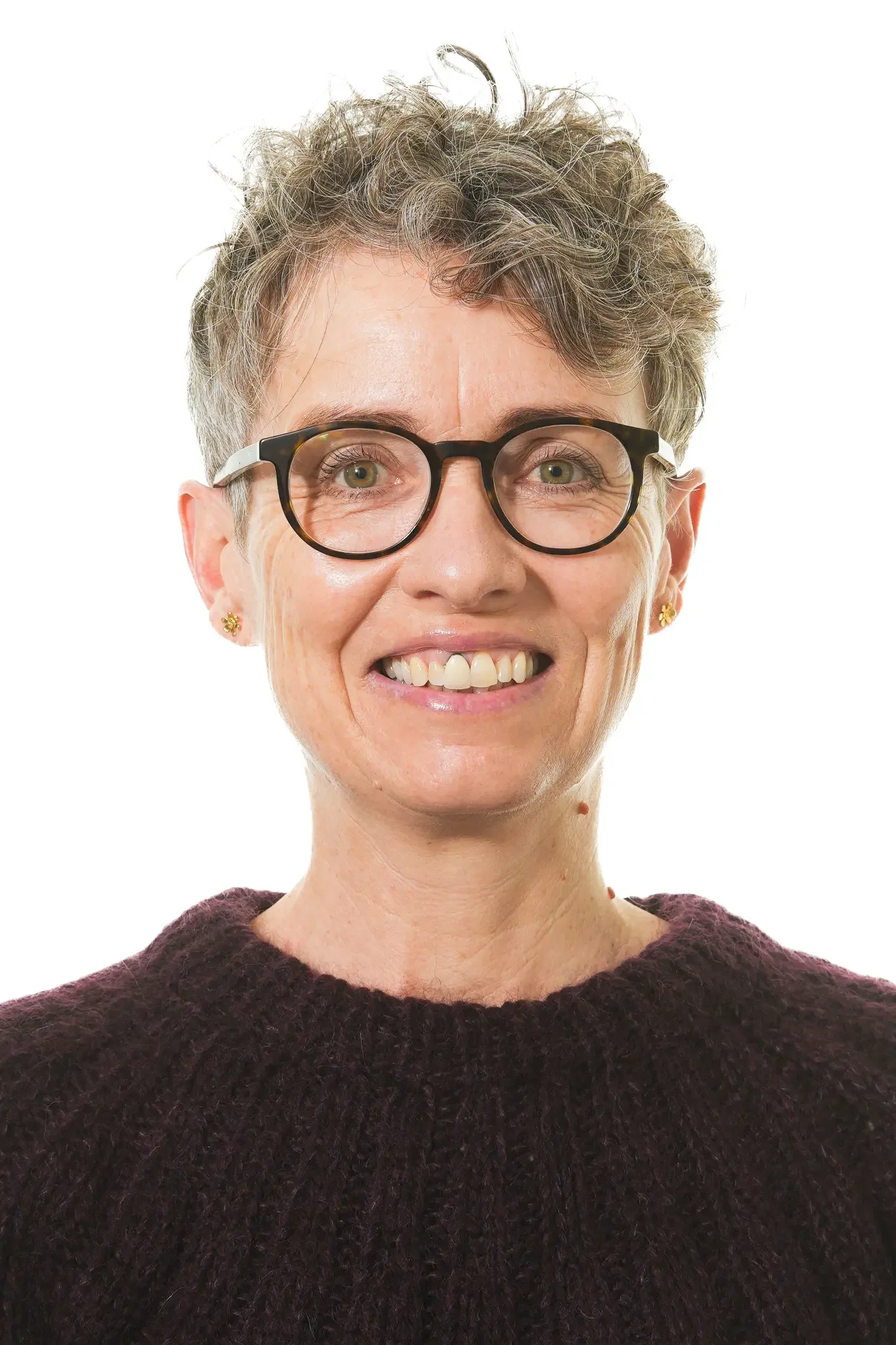 Et smilende kvindemenneske med briller og kort krøllet gråt hår iført en grå skjorte.