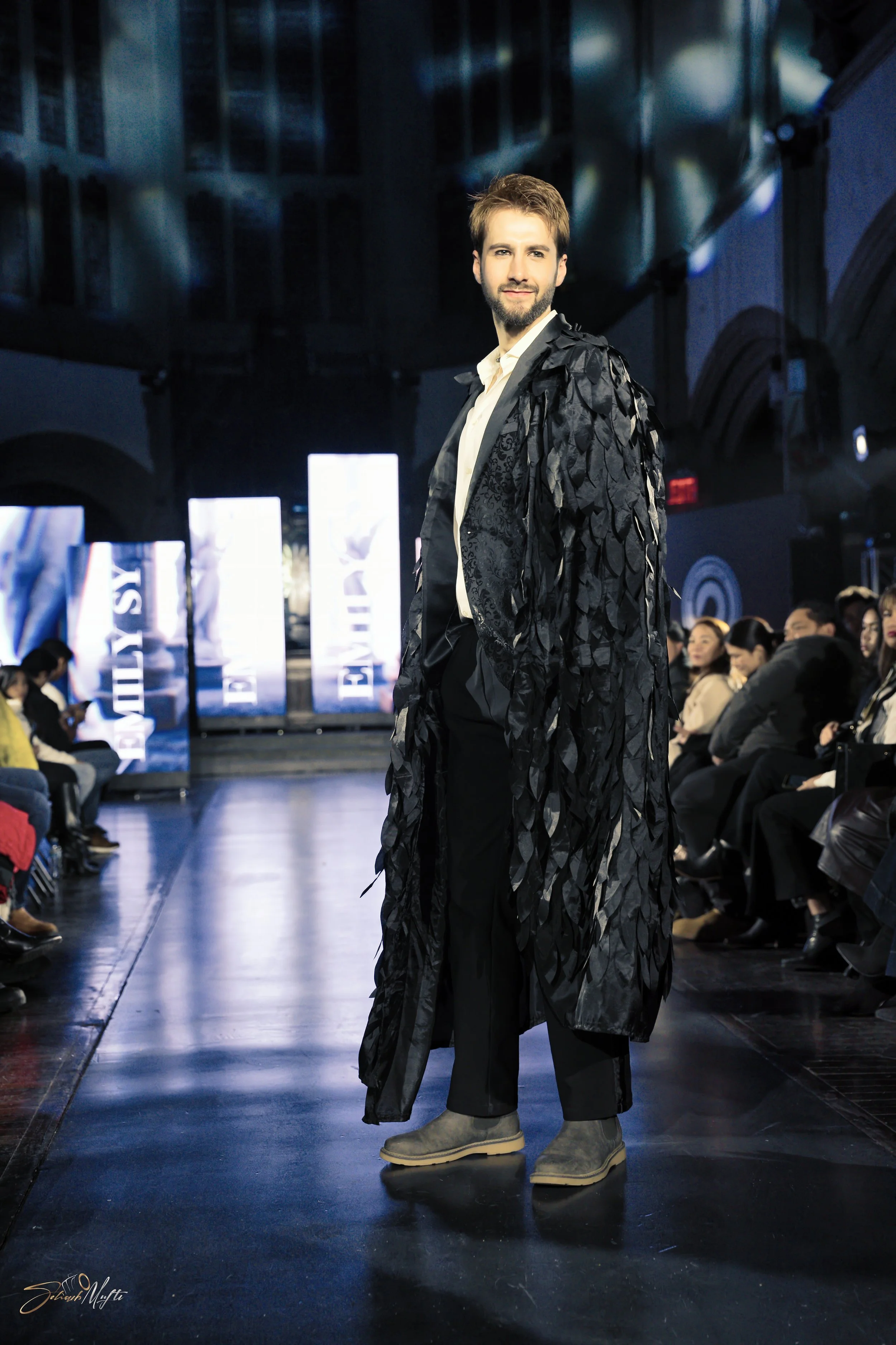 Male model walking on runway at a fashion show, wearing a long black coat with feather-like details, white shirt, black pants, and gray shoes, with audience seated on both sides of the runway.