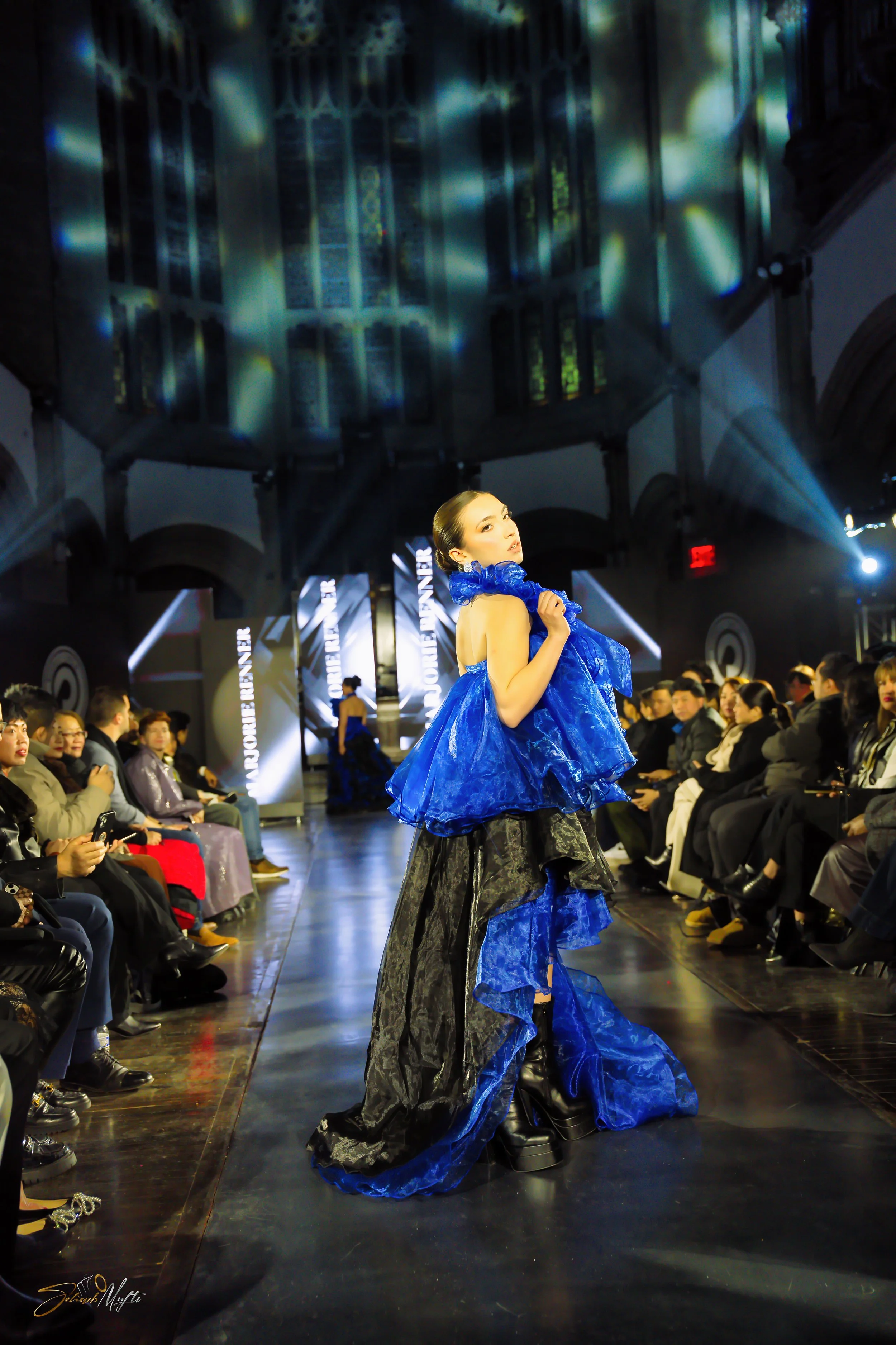 Runway fashion show with model wearing a blue and black gown, audience on either side, in a venue with stained glass windows and dramatic lighting.