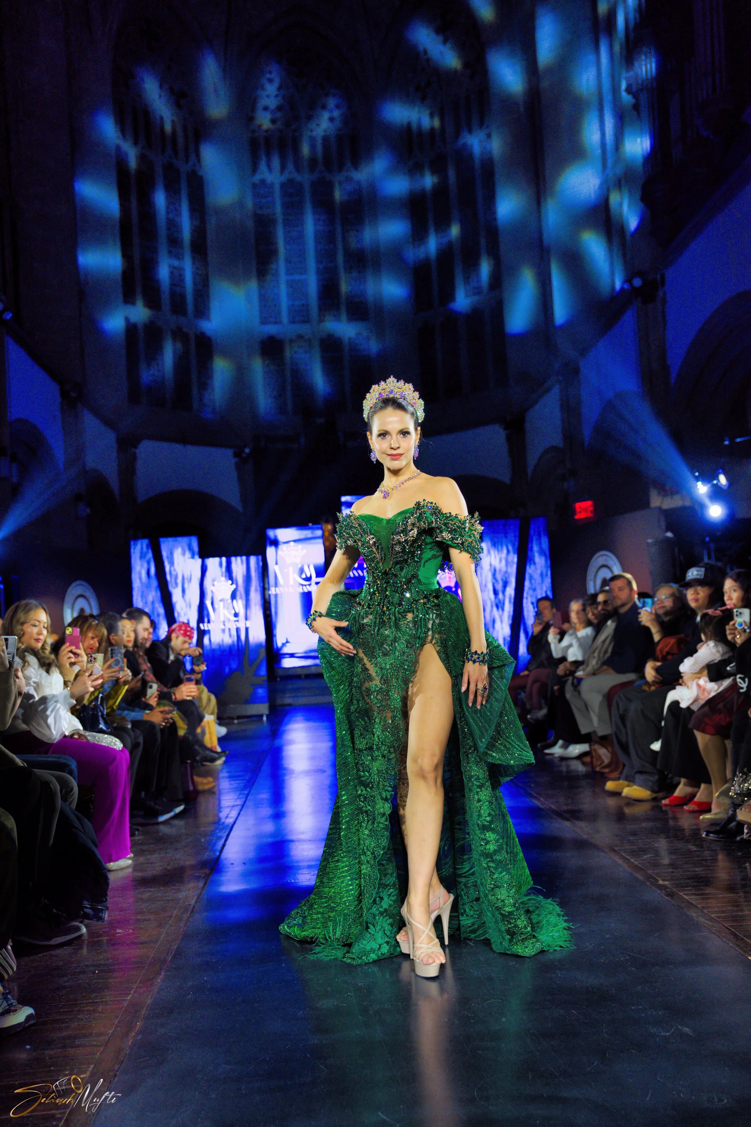 A woman walking runway in a green dress with high slit at a fashion show, audience watching, and stage lights overhead.
