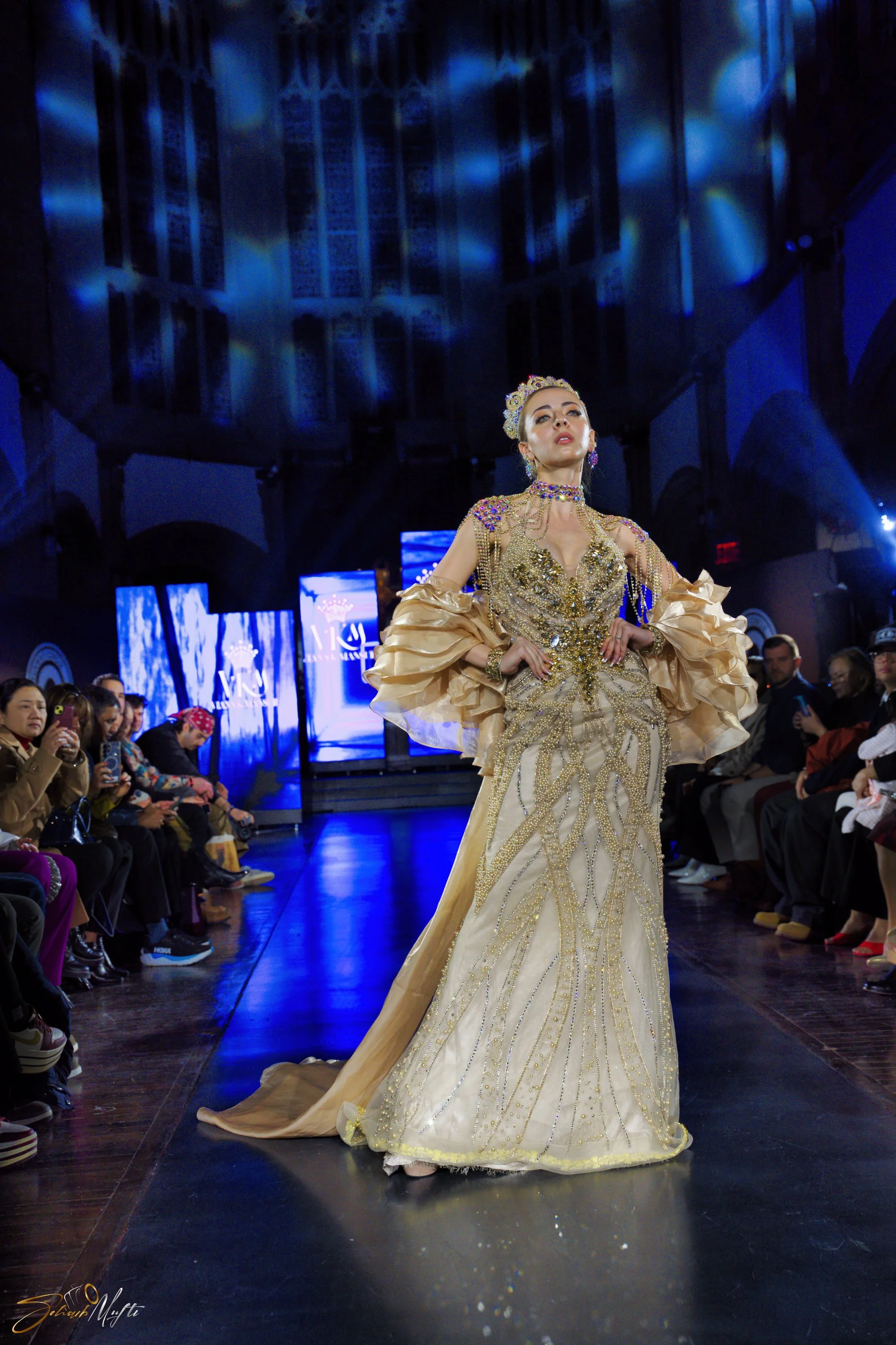 A female model wears an ornate gold gown with intricate embellishments and ruffled sleeves on a runway during a fashion show, with seated audience and digital screens in the background.