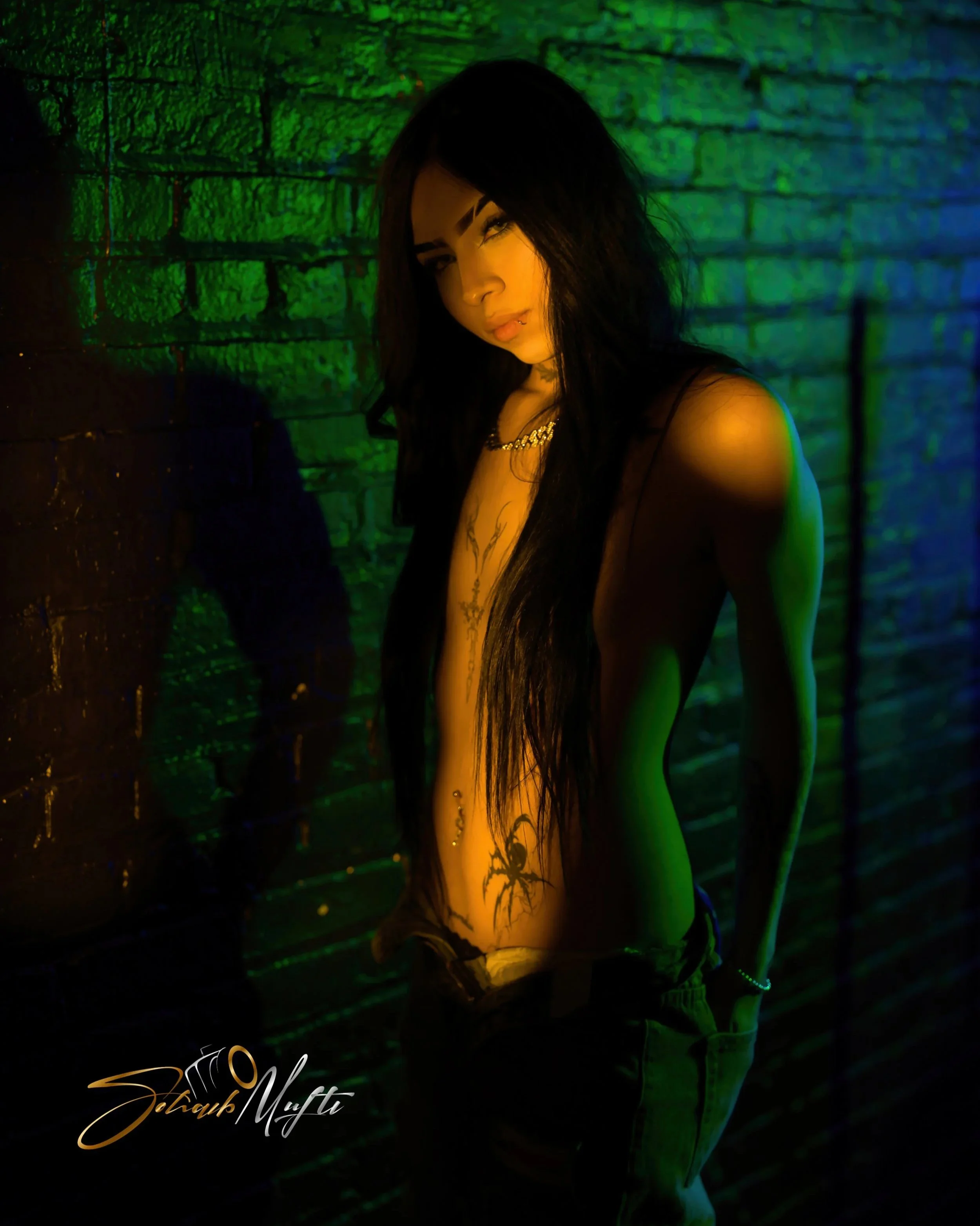 A woman with long black hair stands against a green-lit brick wall, wearing revealing clothing with tattoos on her torso, looking directly at the camera.