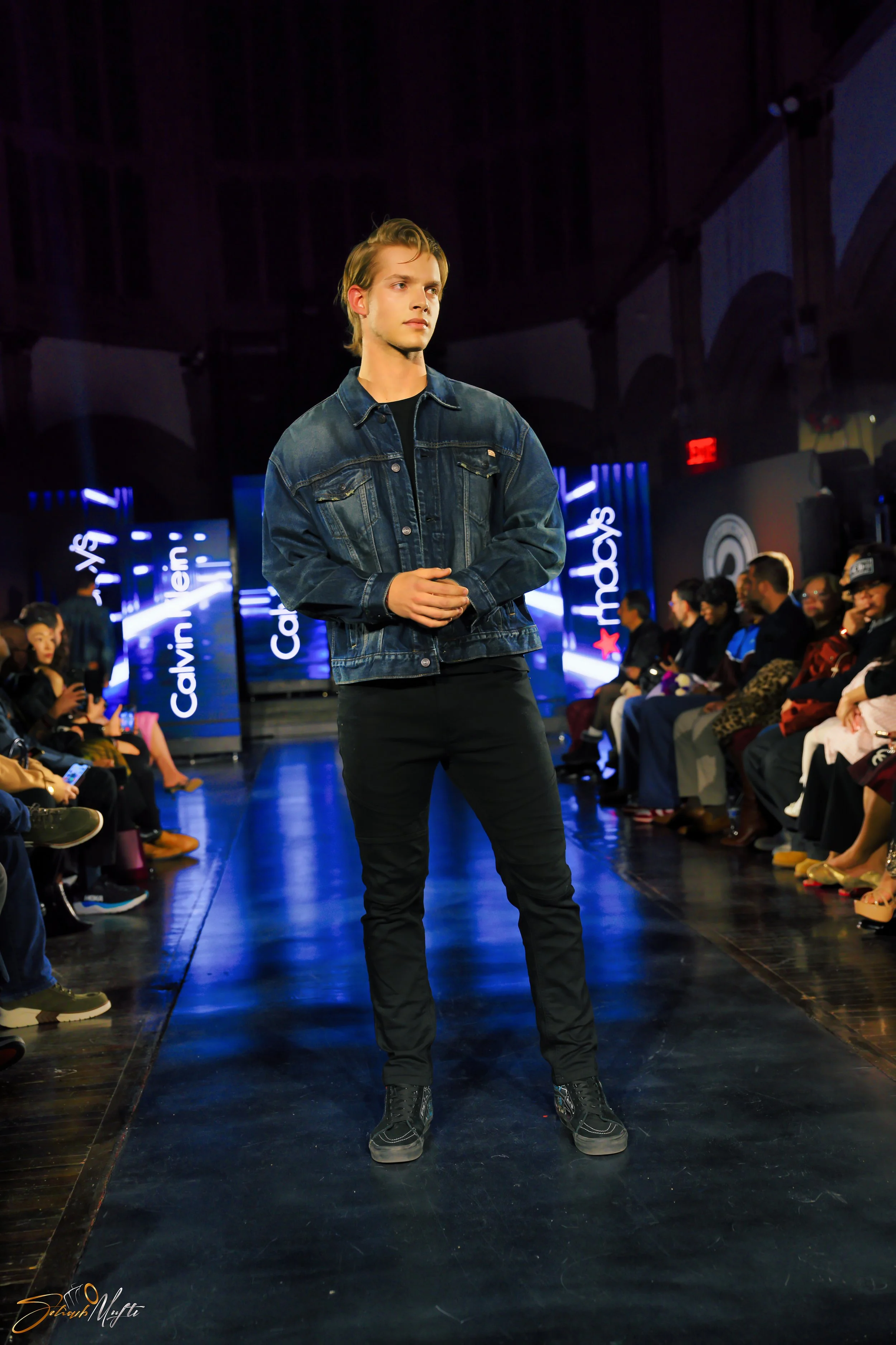 A male model in a denim jacket and black pants walking on a runway at a fashion show with an audience on both sides and bright digital screens in the background.