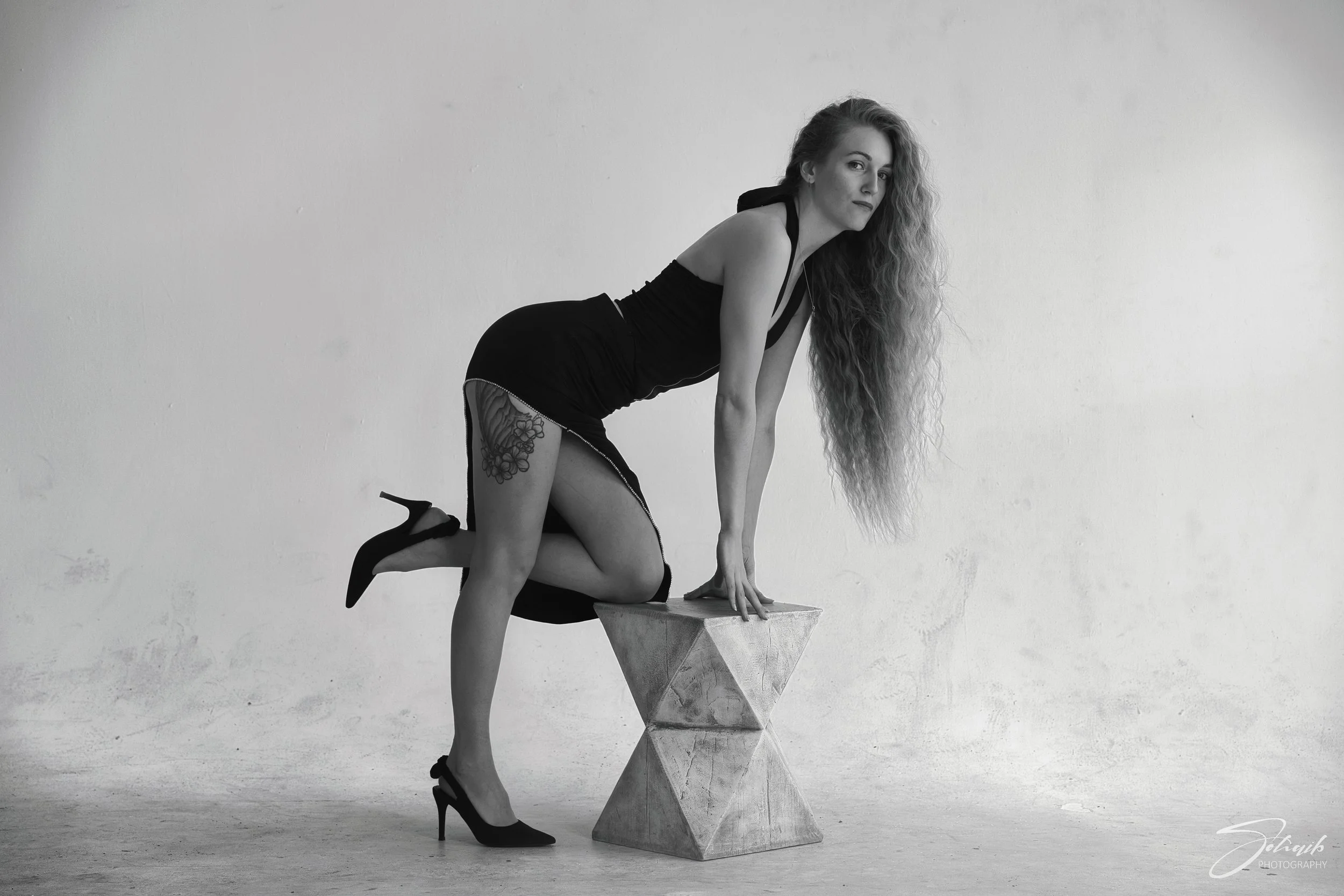 Black and white photo of a woman with long curly hair wearing a sleeveless black dress and high heels, posing with hands and one knee on a geometric block