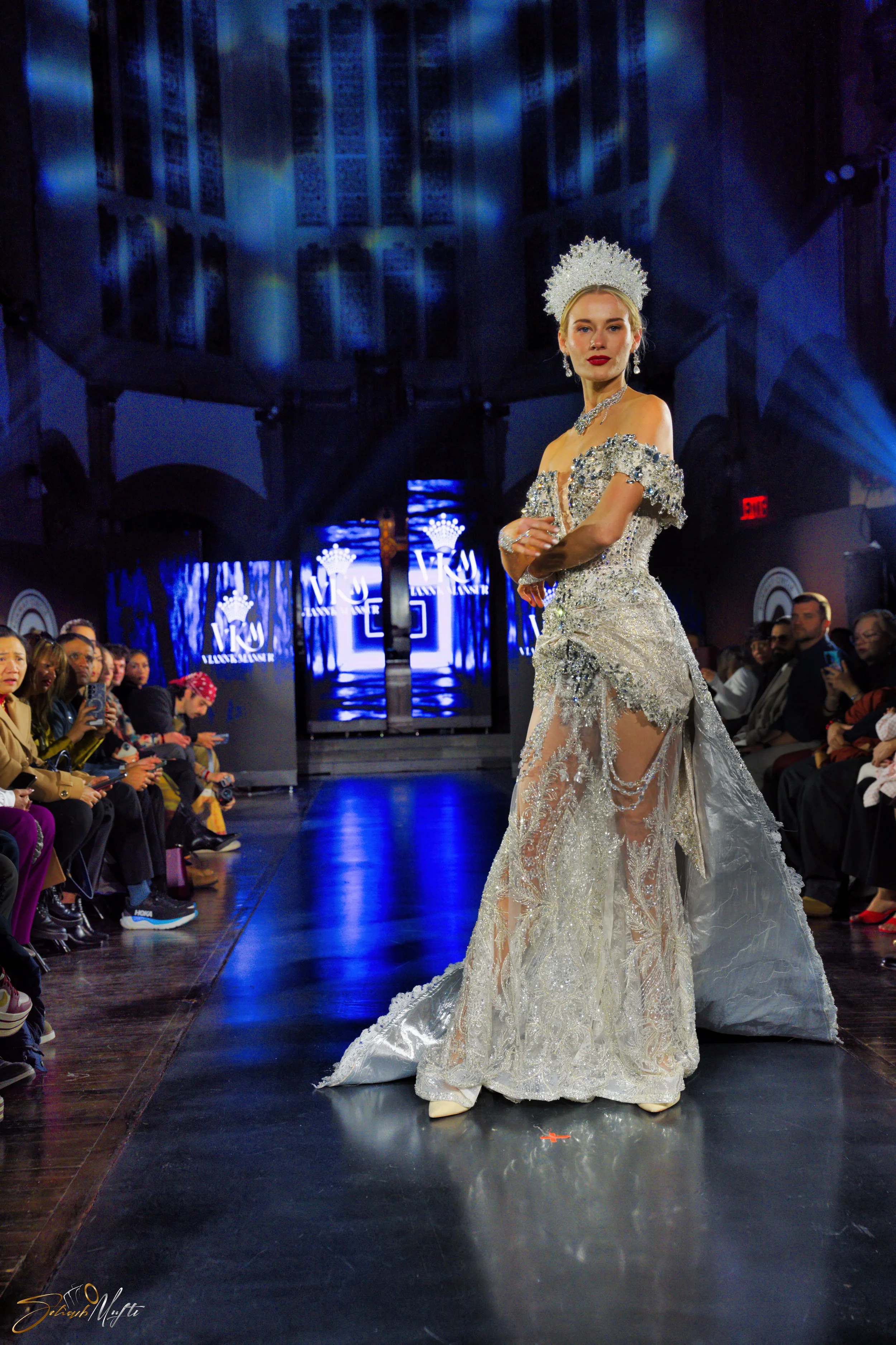 A woman in an ornate, silver, and white gown with embellishments and lace details walks on a runway during a fashion show. She is also wearing a large silver headdress and jewelry, with an audience seated on either side of the runway, watching and ta