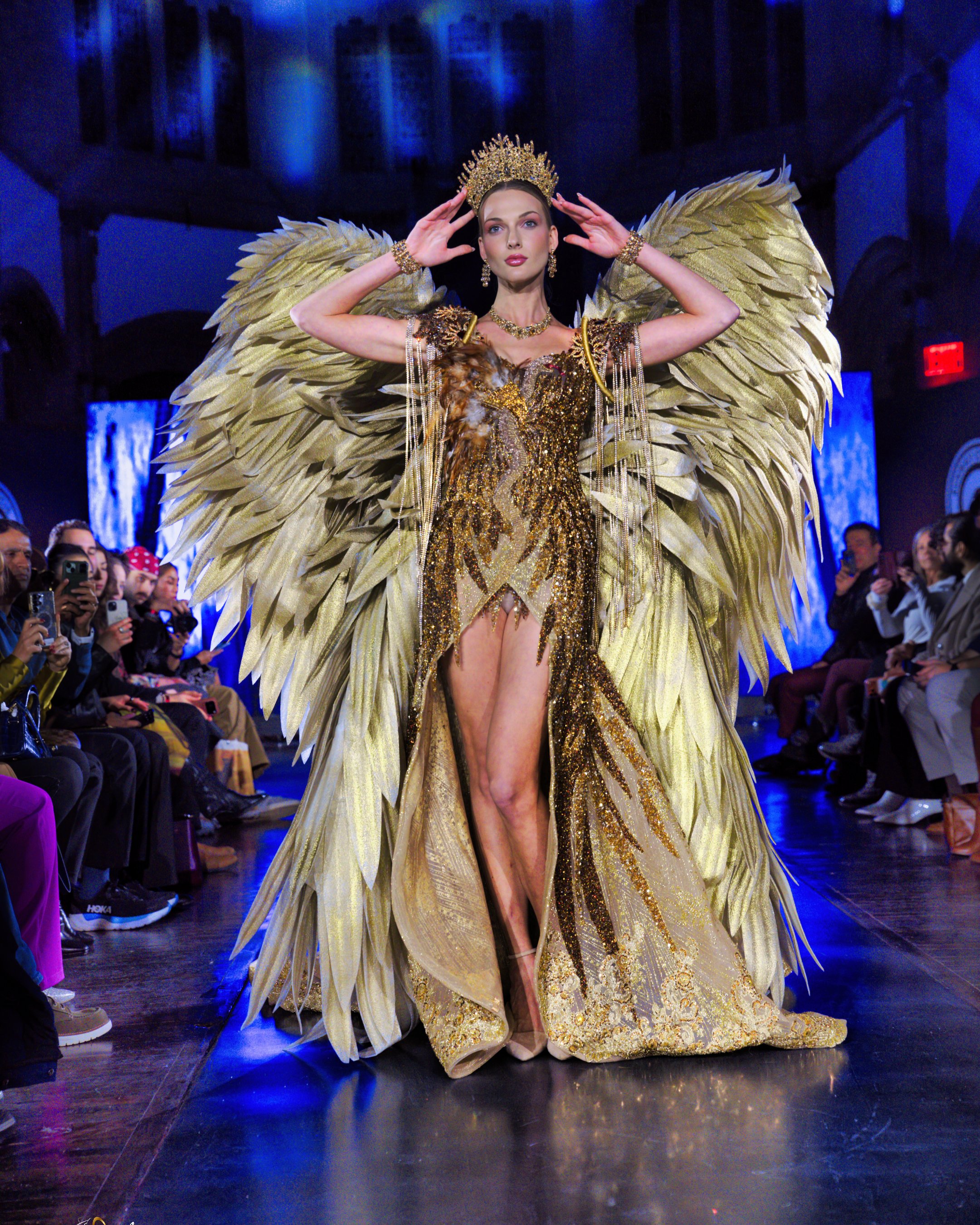 A woman on a runway dressed in an elaborate gold costume with large feathered wings and a crown, surrounded by audience members taking photos.