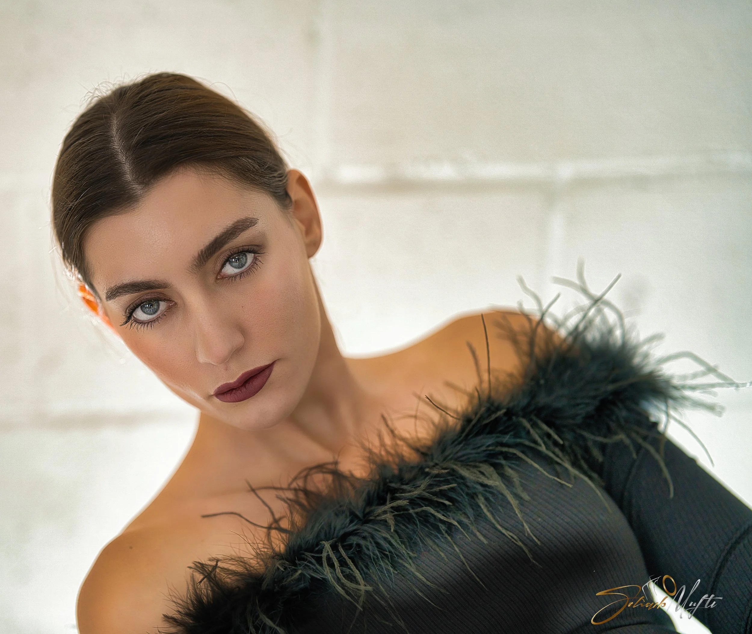 A woman with short brown hair and striking blue eyes, wearing dark lipstick, a black off-the-shoulder dress with feathered detailing, posed against a light background.