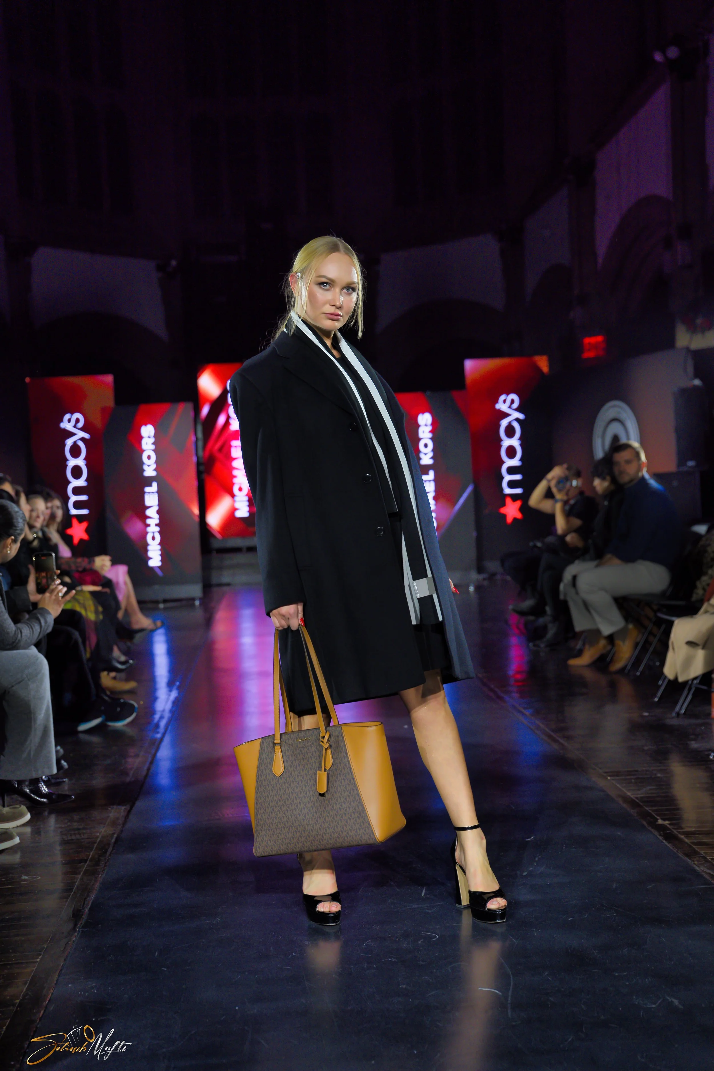 A woman walking on a runway at a fashion show, carrying a brown designer handbag, dressed in a black coat, high heels, and a striped dress, with a digital screen behind her displaying the "Macy's" and "Michael Kors" logos.