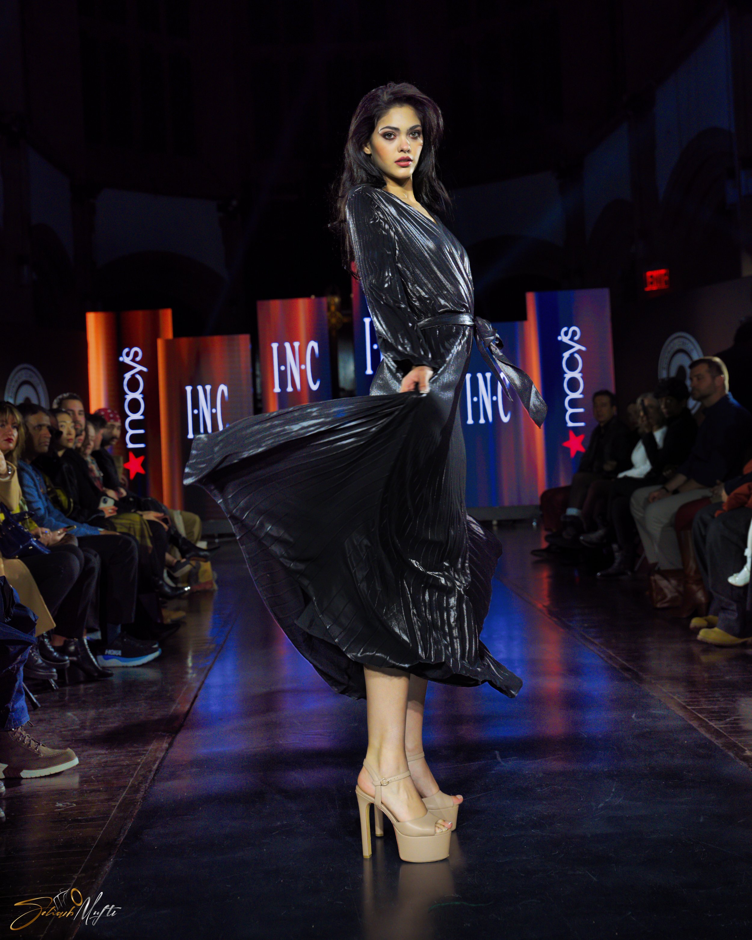 A female model on a runway at Macy's, wearing a black shiny pleated dress with beige high heels, holding the edge of her dress, surrounded by an audience and Macy's signage in the background.