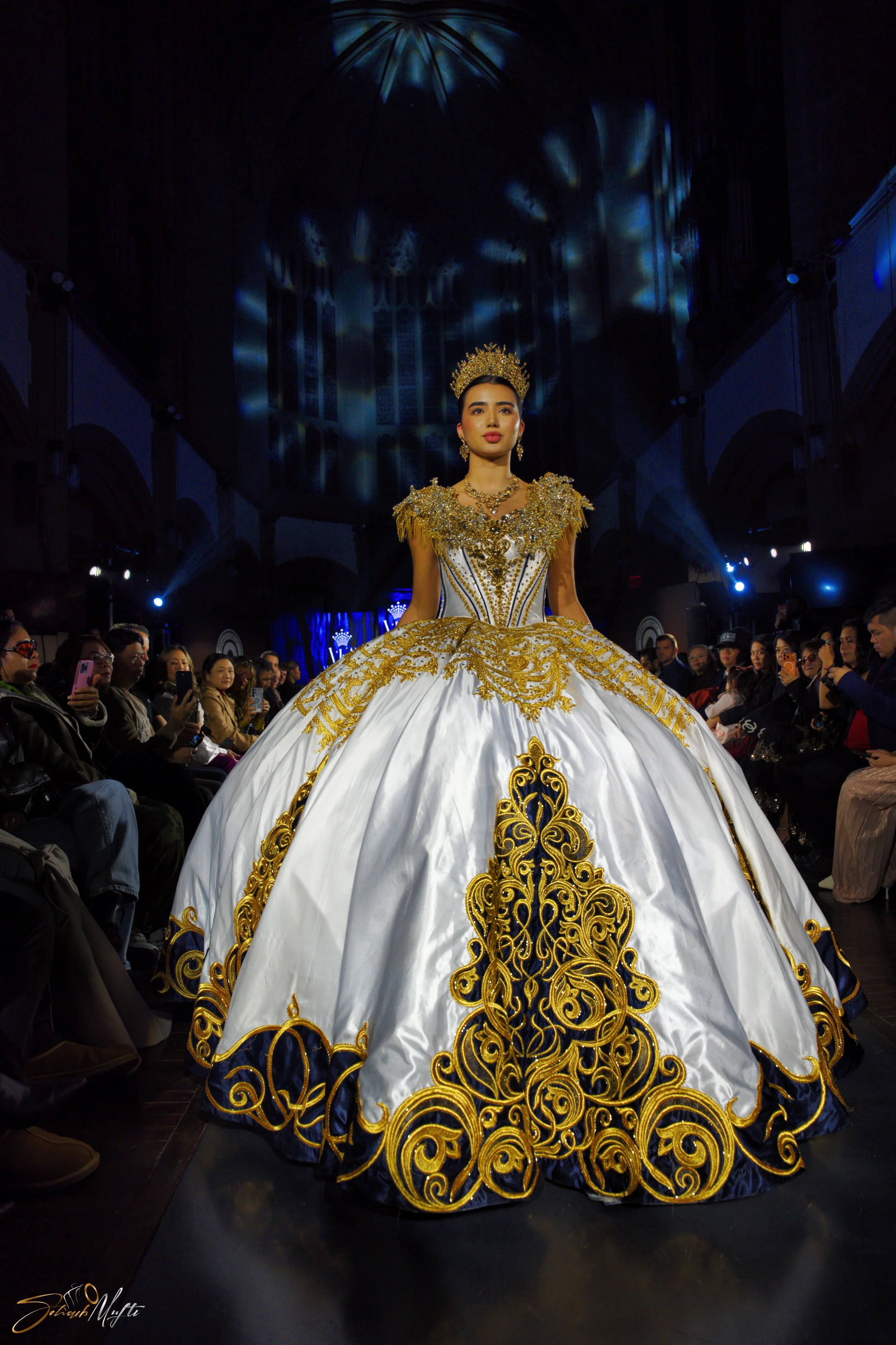A woman in a white and gold ornate gown and crown walking on a runway during a fashion show.