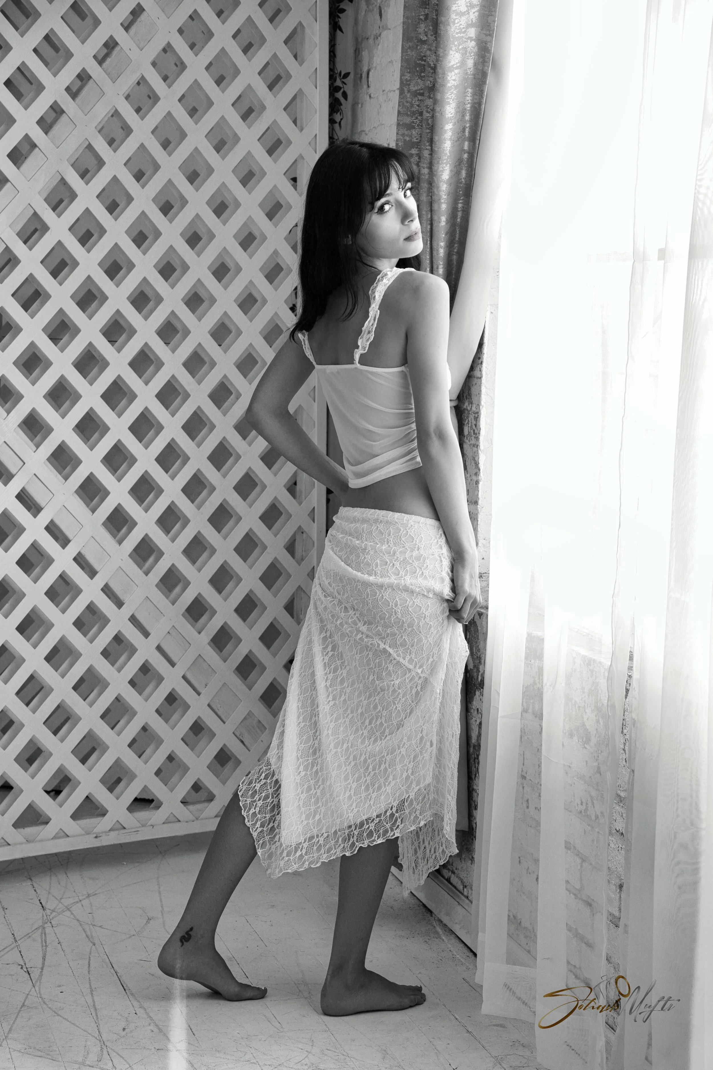 A woman with dark hair in lingerie, standing barefoot near a window with sheer curtains, looking over her shoulder at the camera, with a lace skirt, in a monochrome photograph.