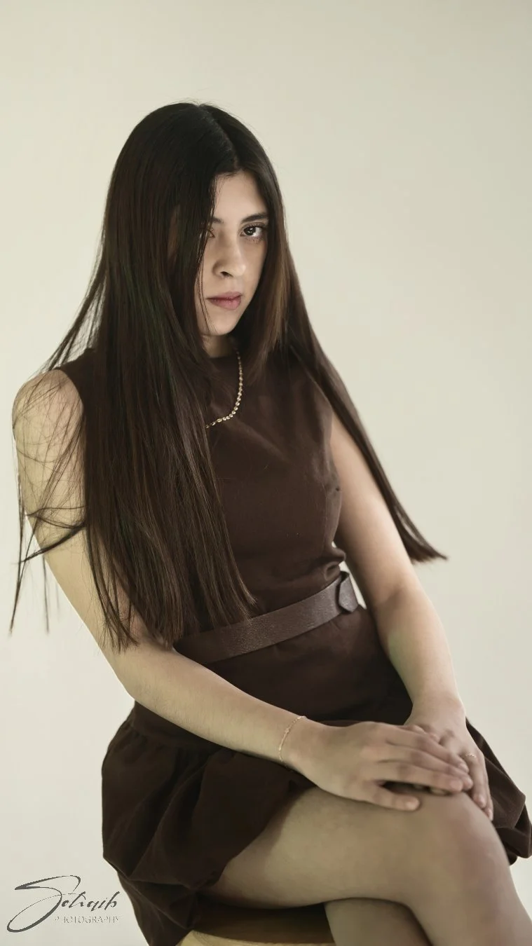 A woman with long dark hair wearing a brown dress and sitting on a chair, looking at the camera with a serious expression.