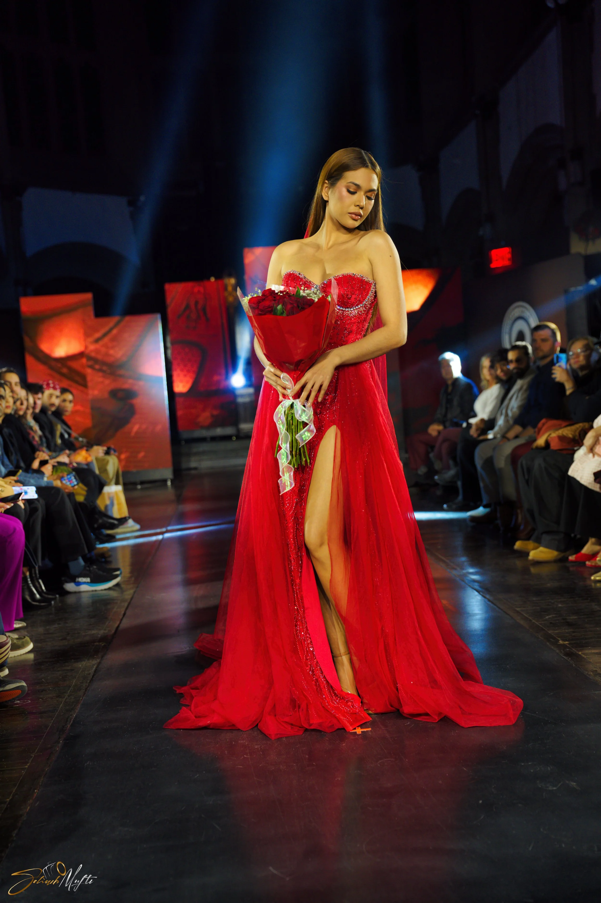 A woman in a red gown with a high slit walks the runway while holding a bouquet of flowers, surrounded by seated spectators at a fashion show.