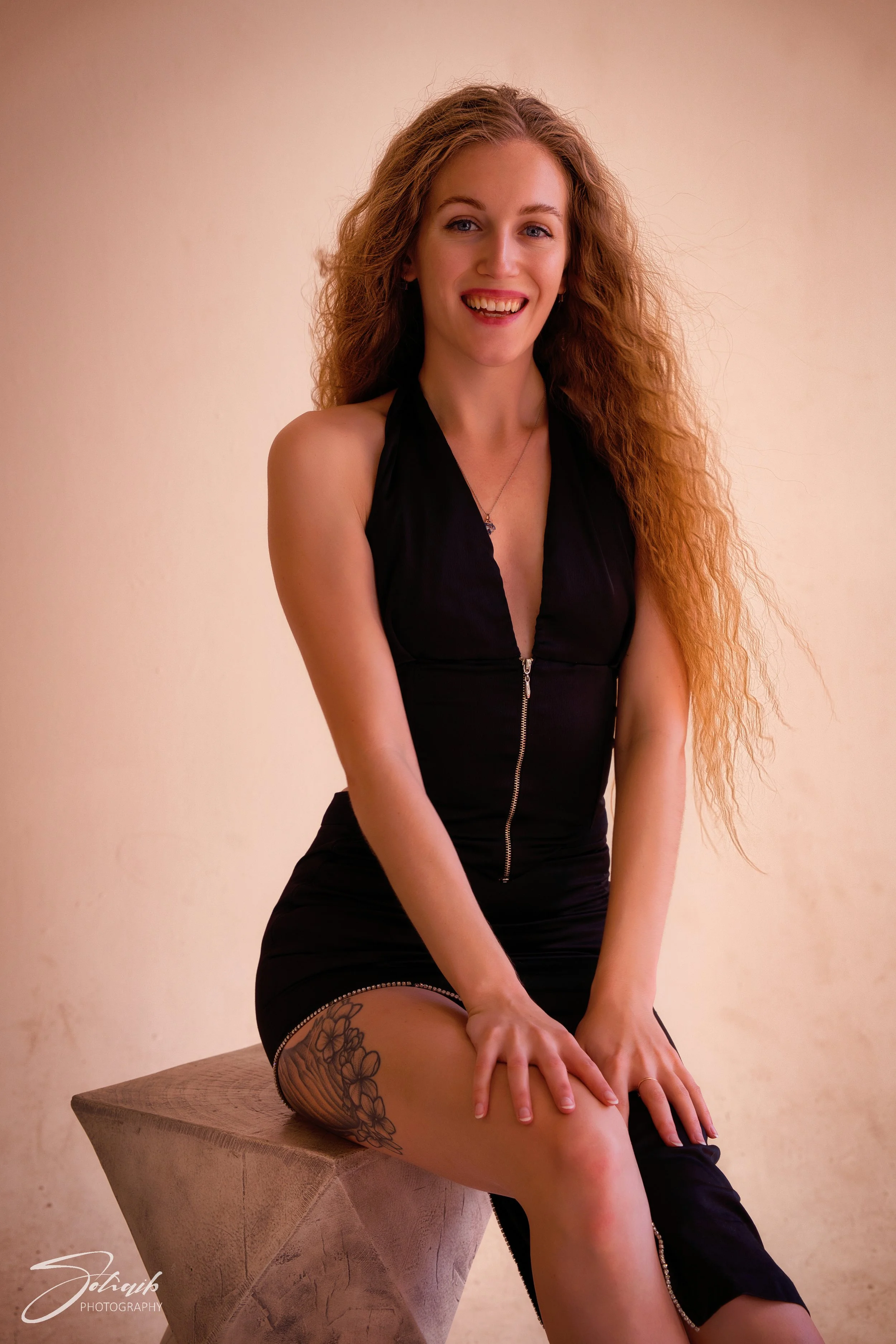 A young woman with long, curly red hair sitting on a wooden stool, smiling at the camera, wearing a black sleeveless dress with a zipper in the front and a floral tattoo on her left thigh.