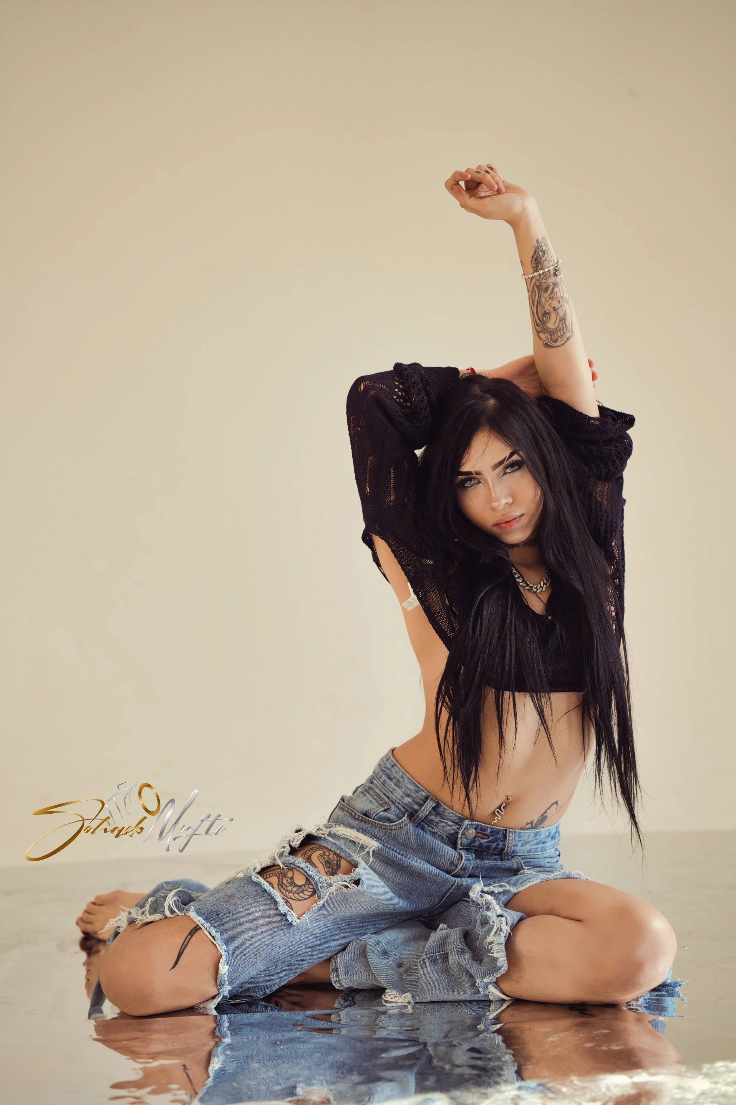 A woman with long black hair, wearing a black crop top and ripped jeans, poses sitting on the floor with her knees bent and arms raised overhead, in front of a plain beige background.