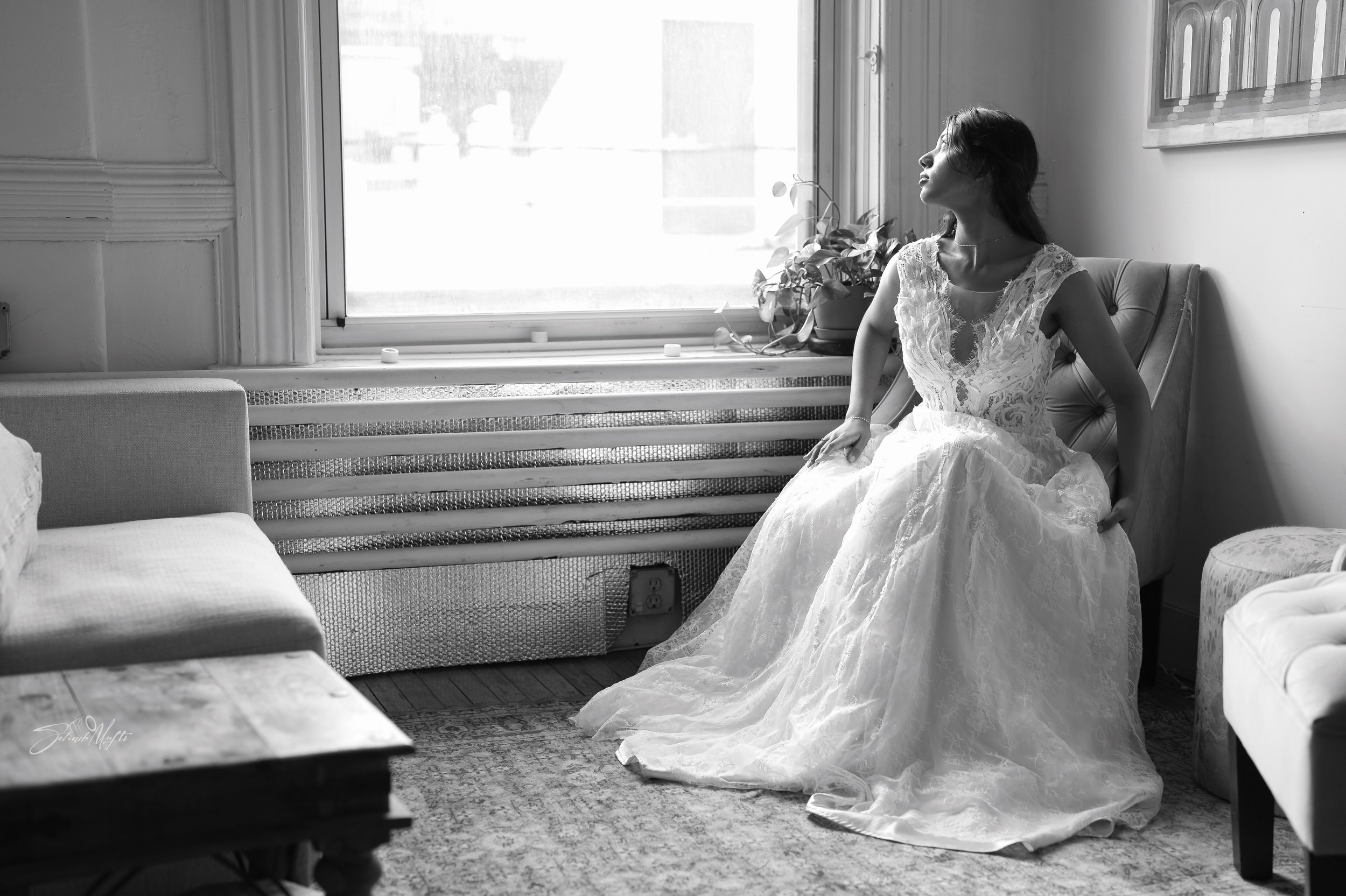 A woman in a wedding dress sitting on a chair by a window, looking upwards with sunlight coming through the window.