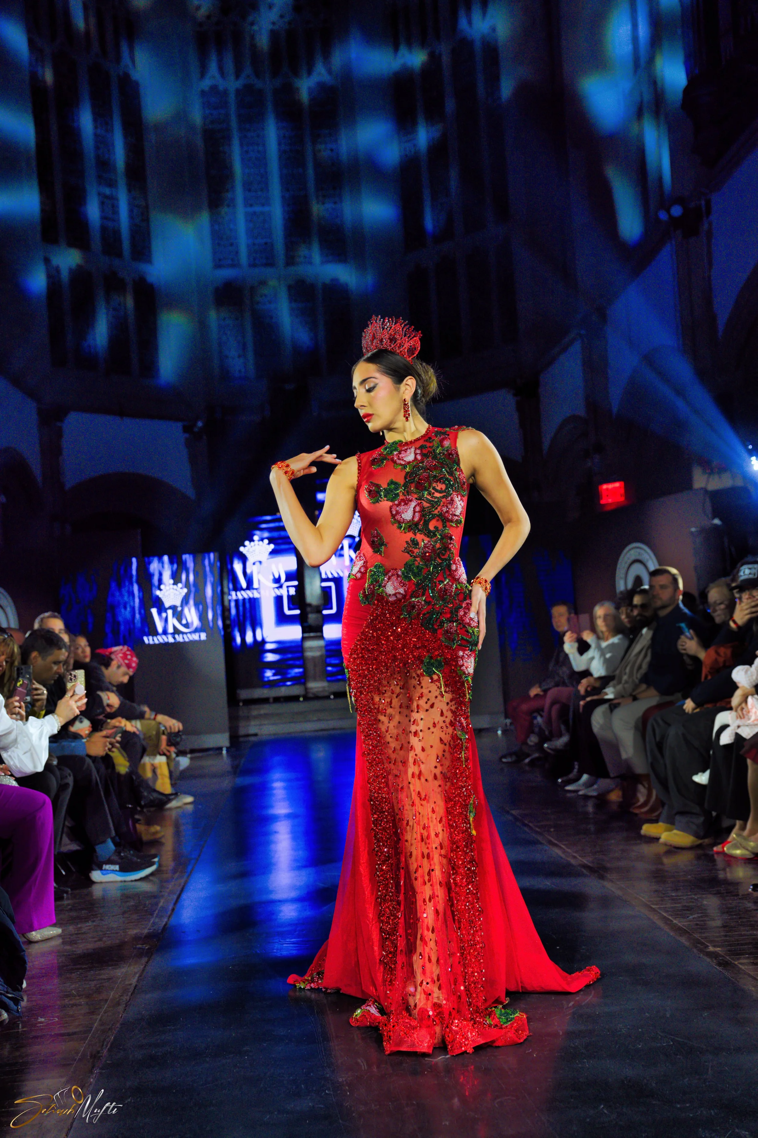 A woman in a red, embroidered, sheer gown walks on a fashion runway during a show, with an audience watching on either side and bright stage lighting overhead.
