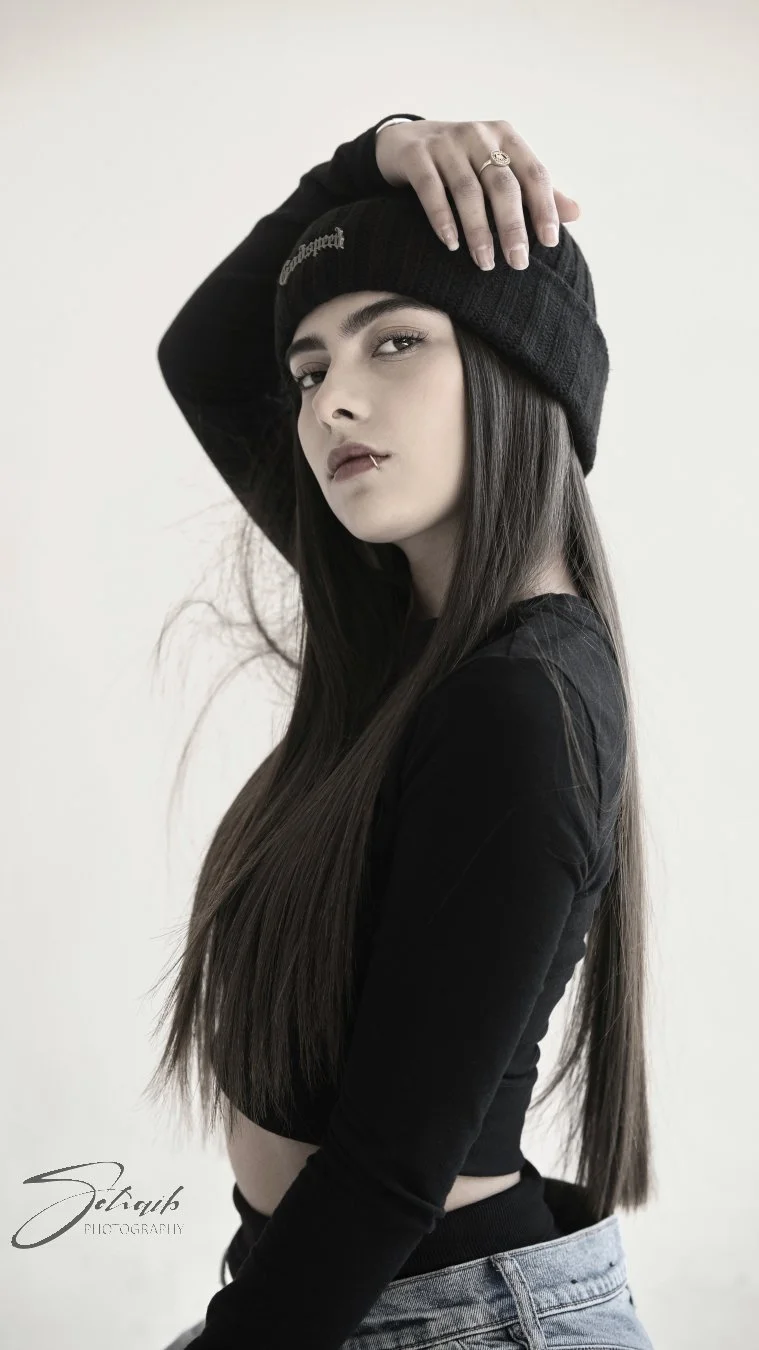 Young woman with long dark hair wearing a black beanie hat, black long-sleeve top, and high-waisted jeans, posing indoors against a plain background.