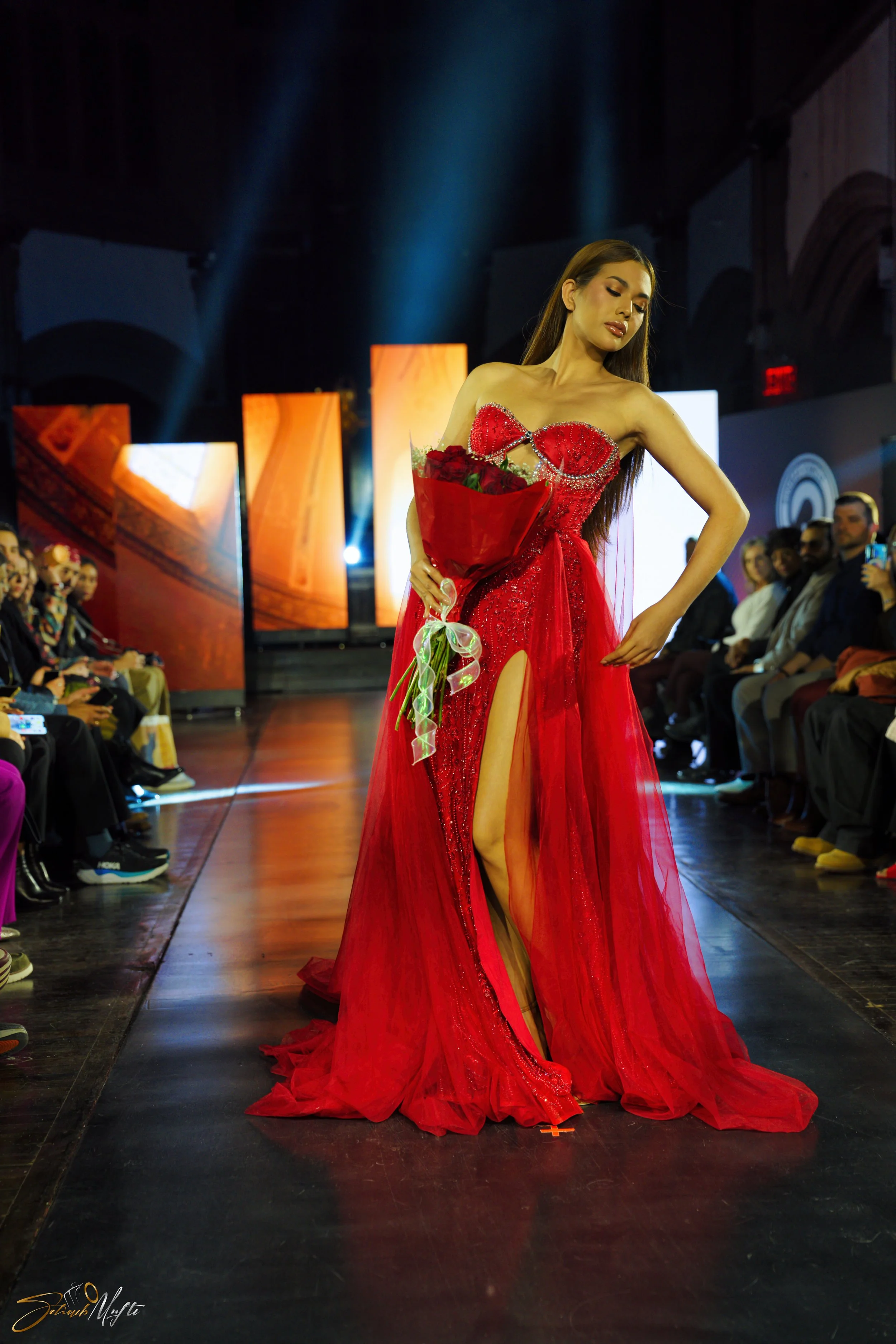 A woman walking on a fashion runway wearing a red gown with a thigh-high slit, holding a bouquet of red roses, with an audience watching in the background.