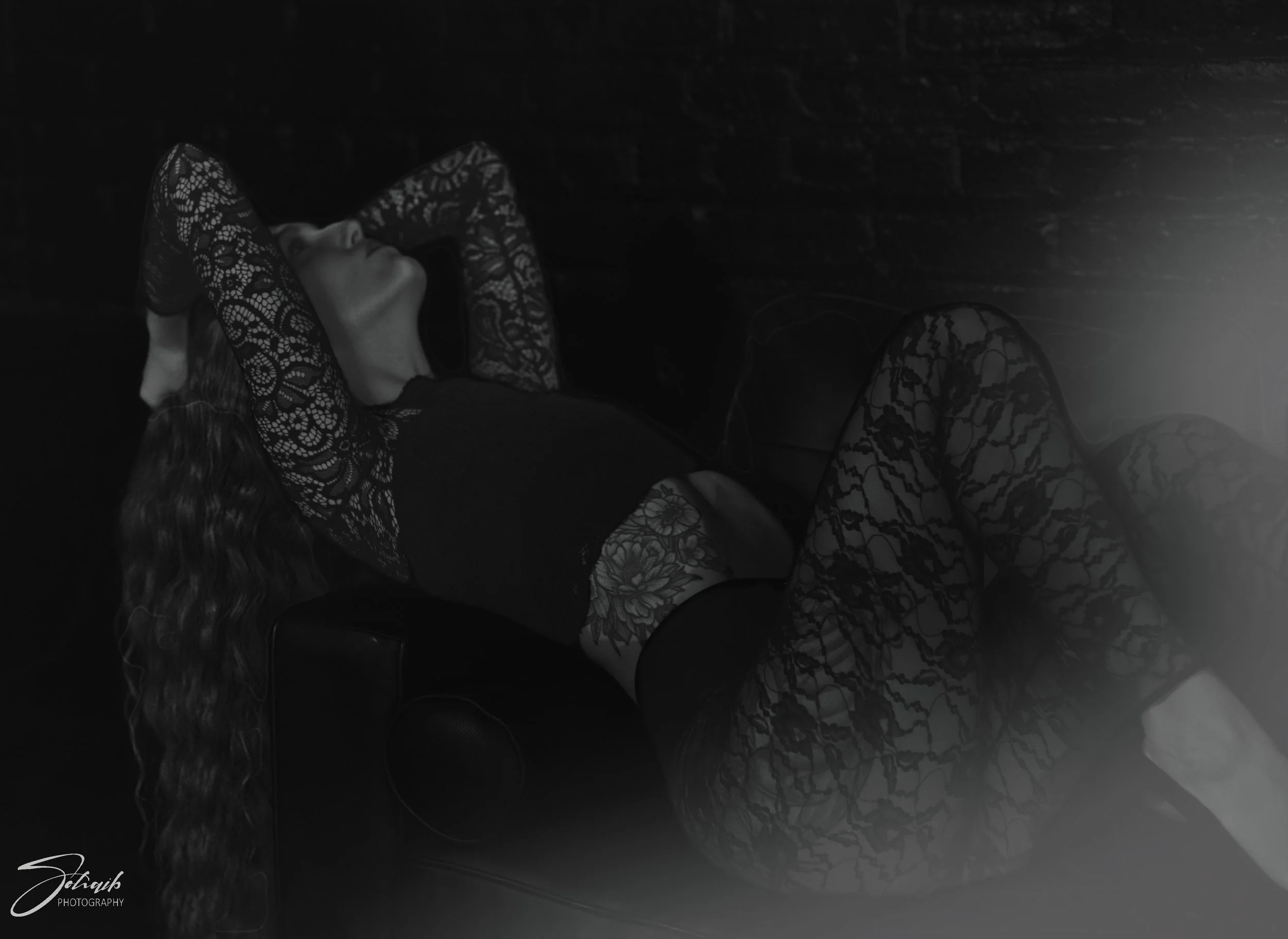 A woman with long, curly hair lying on a couch with her eyes closed, wearing a black top and lace-patterned sheer sleeves and pants, in a dark room with a brick wall background.