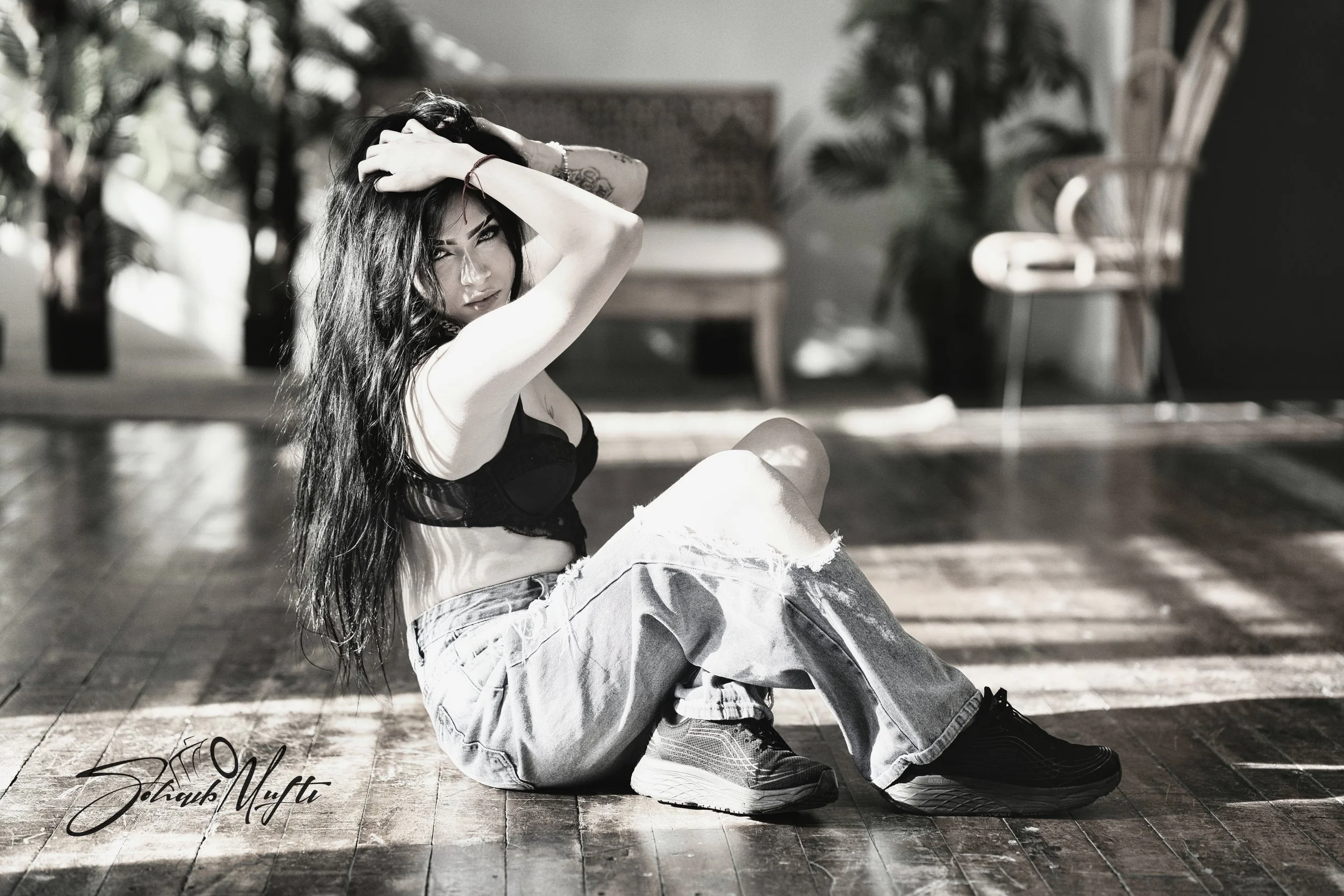 A woman with long dark hair wearing a black top, ripped jeans, and sneakers, sitting on a wooden floor in a room with large windows and indoor plants, posing with her hands on her head and looking at the camera.