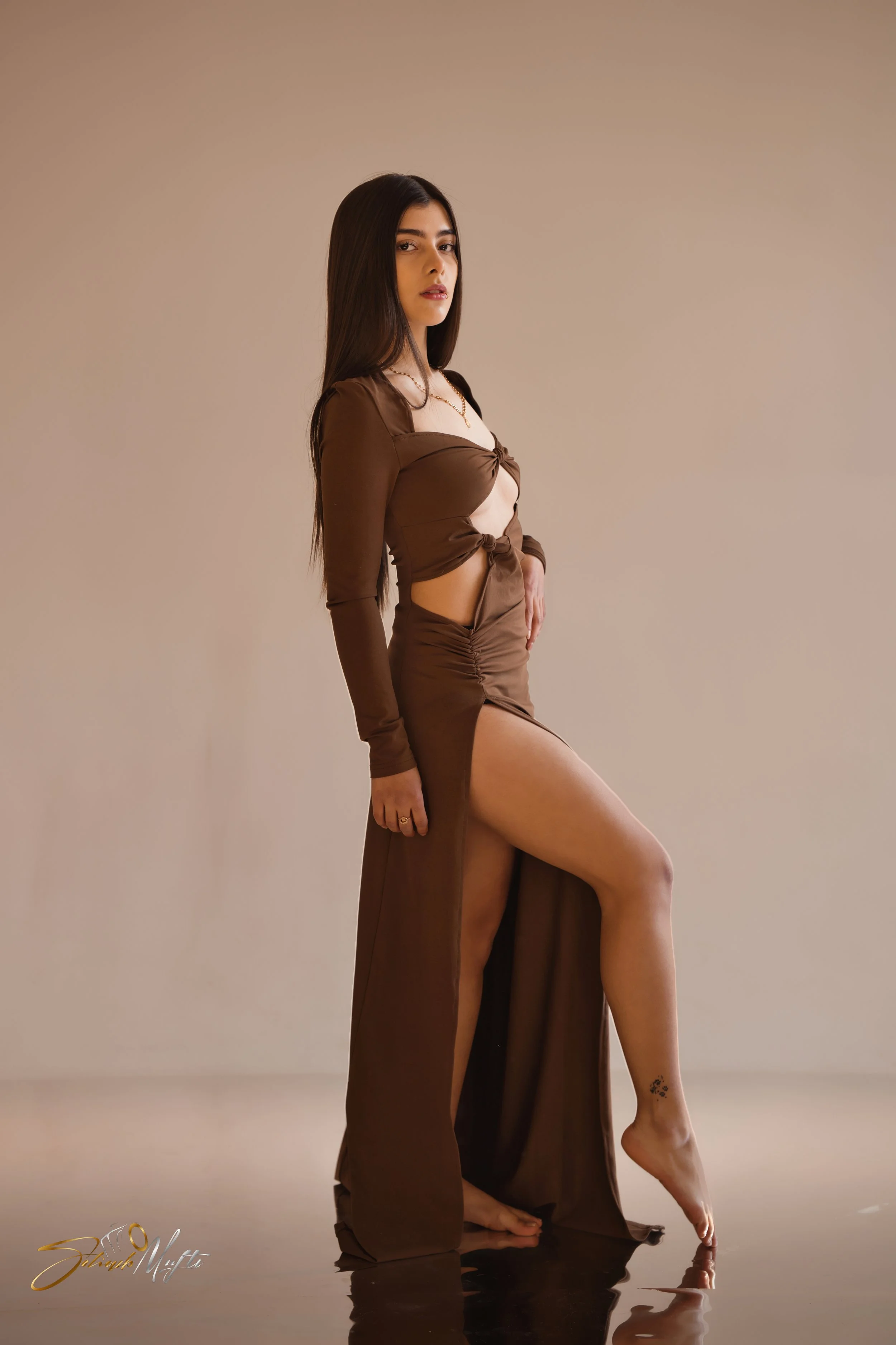 A young woman with long dark hair poses against a plain light beige background. She is wearing a brown, long-sleeved, tied crop top, and matching high-slit skirt. She is standing with one leg extended forward, showing a tattoo on her ankle, and is ba