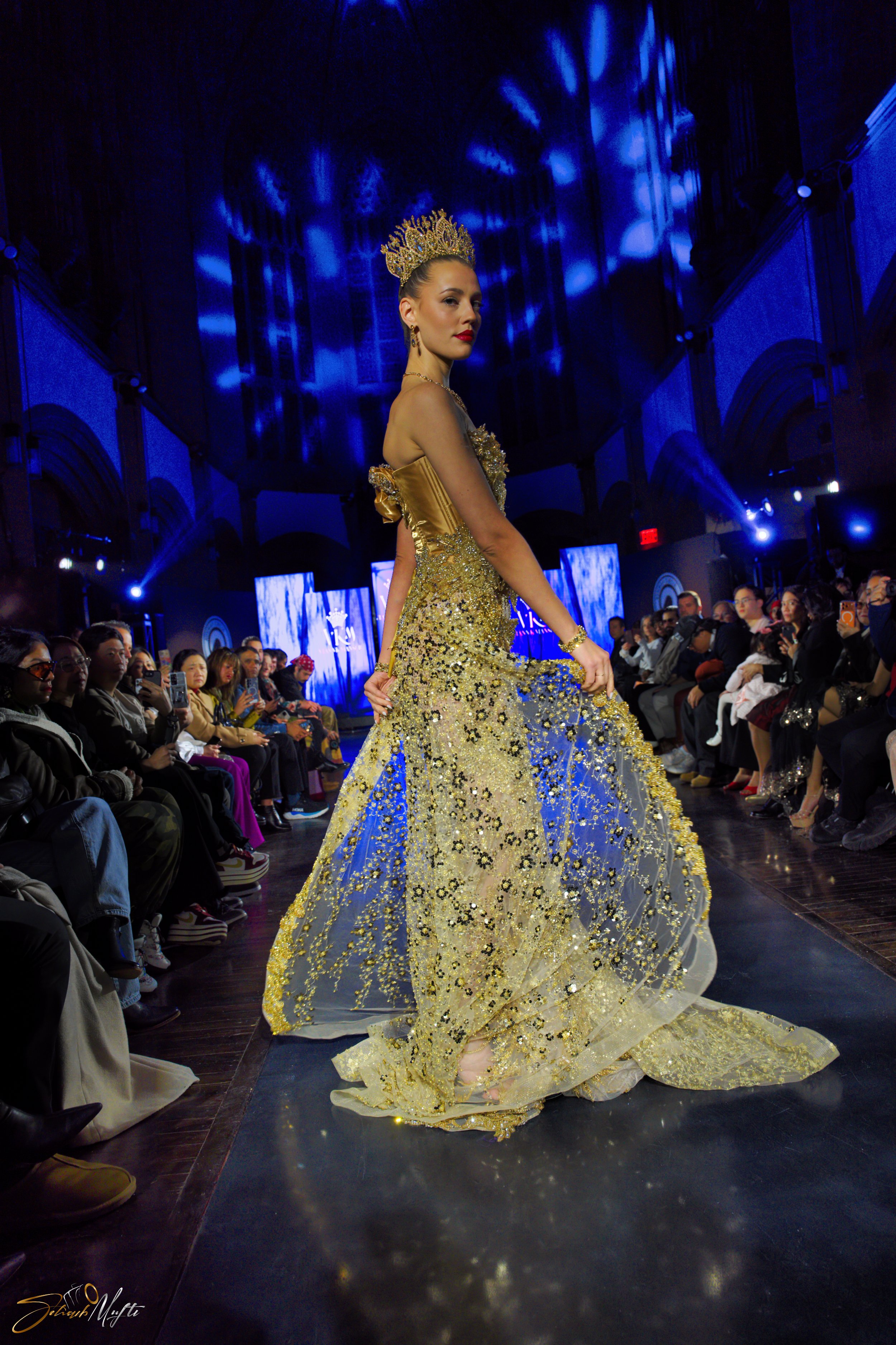A model walking on the runway during a fashion show, wearing a gold and transparent embellished gown with a crown and jewelry, with audience seated on either side and blue stage lighting.