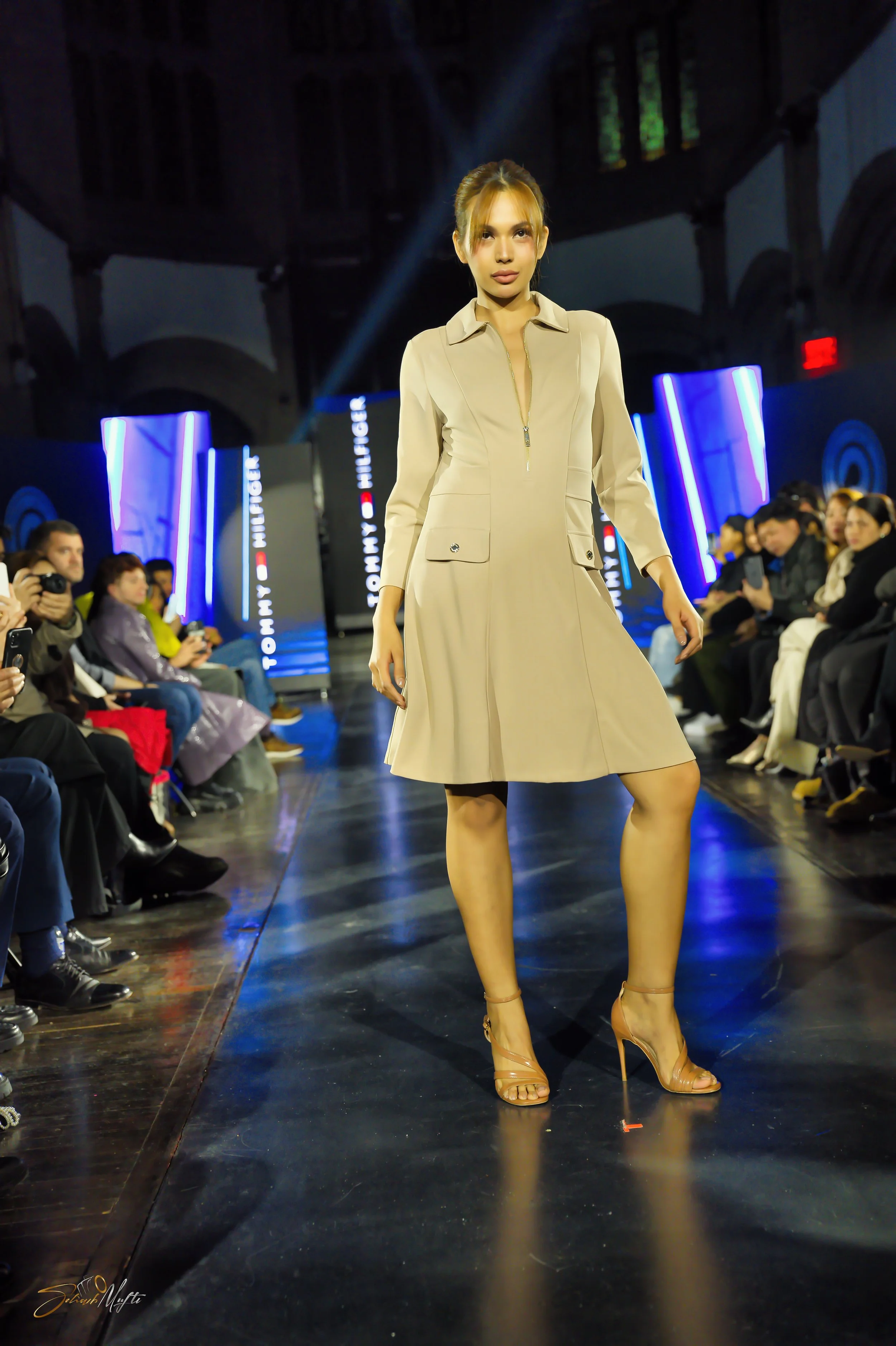 A female model walking on a runway during a fashion show, wearing a beige dress with a zipper front and high heels, with an audience seated on either side of the runway.