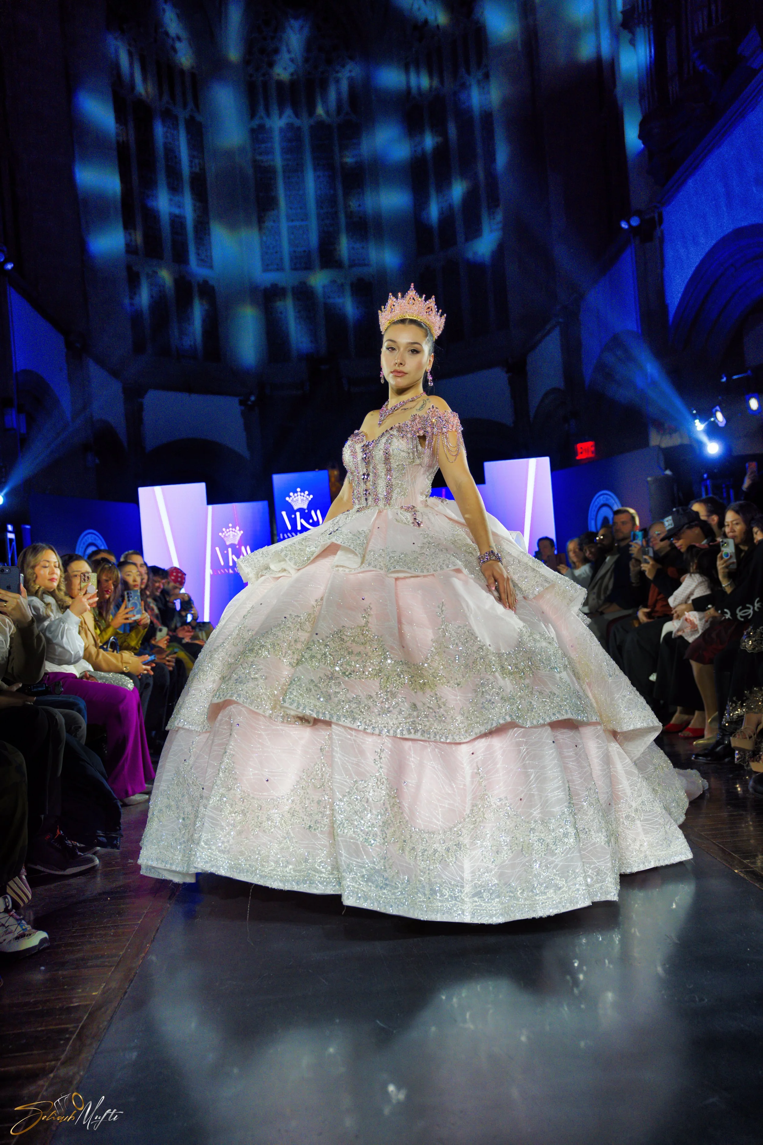 Model on runway wearing an elaborate pink and silver ball gown with a tiara, surrounded by seated audience in a dark, ornately decorated venue with stage lighting.