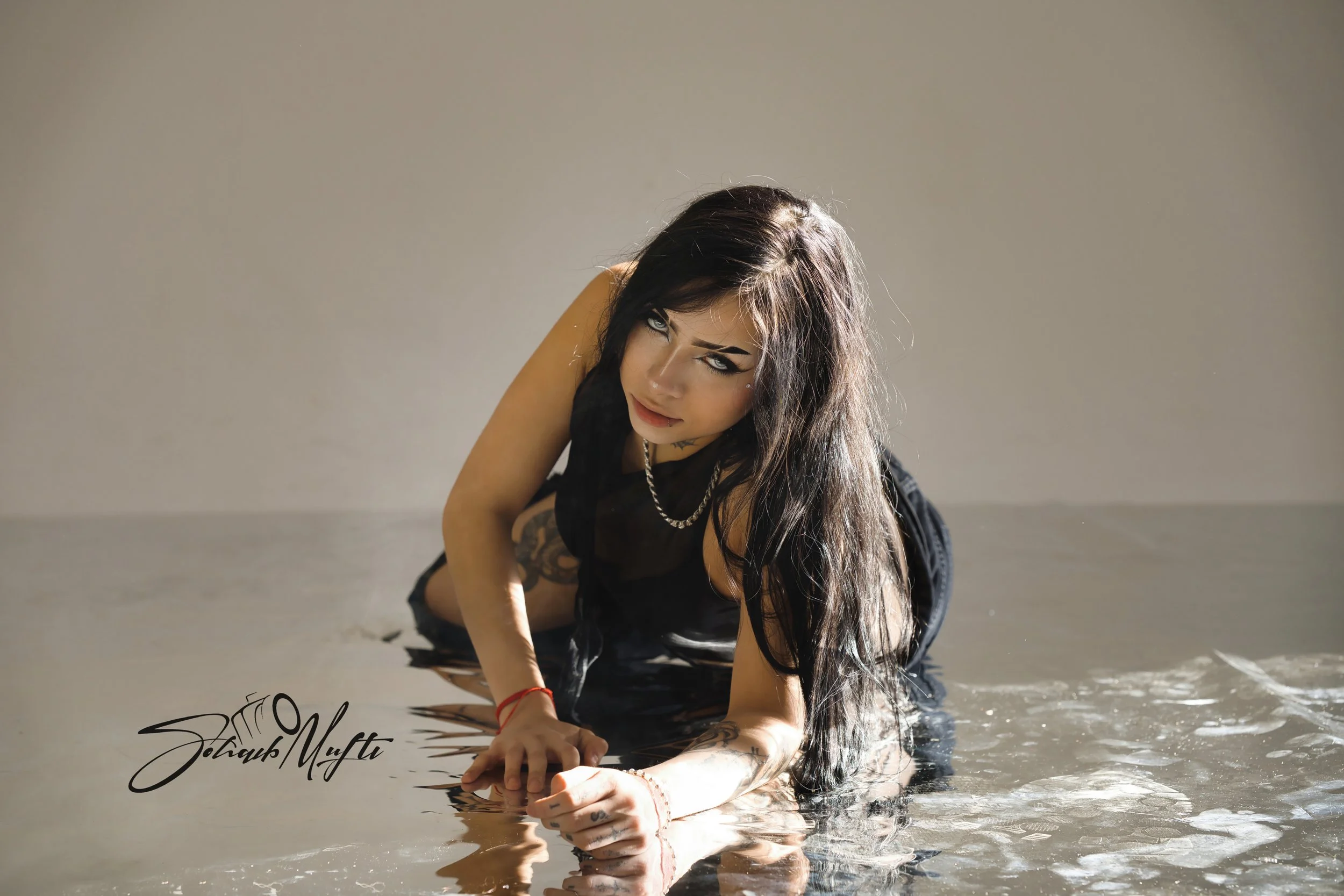 A woman with long dark hair and tattoos on her arm, wearing a black top, crouches on the wet ground with her hands in water, looking up at the camera with a serious expression.