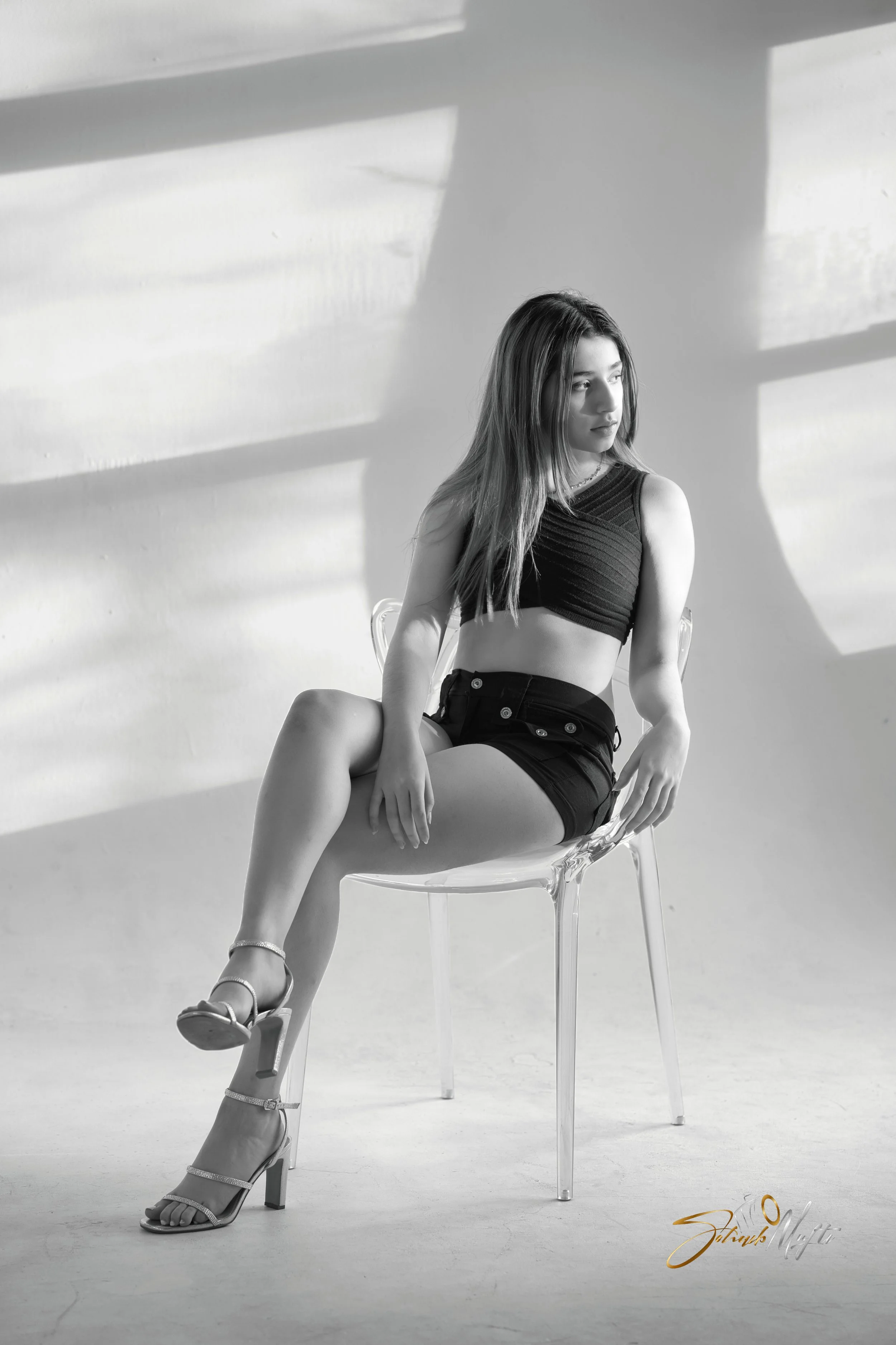 Black and white photo of a young woman sitting on a transparent chair, wearing a black crop top and shorts, with high-heeled sandals, looking away from the camera, with sunlight casting shadow patterns on the wall behind her.