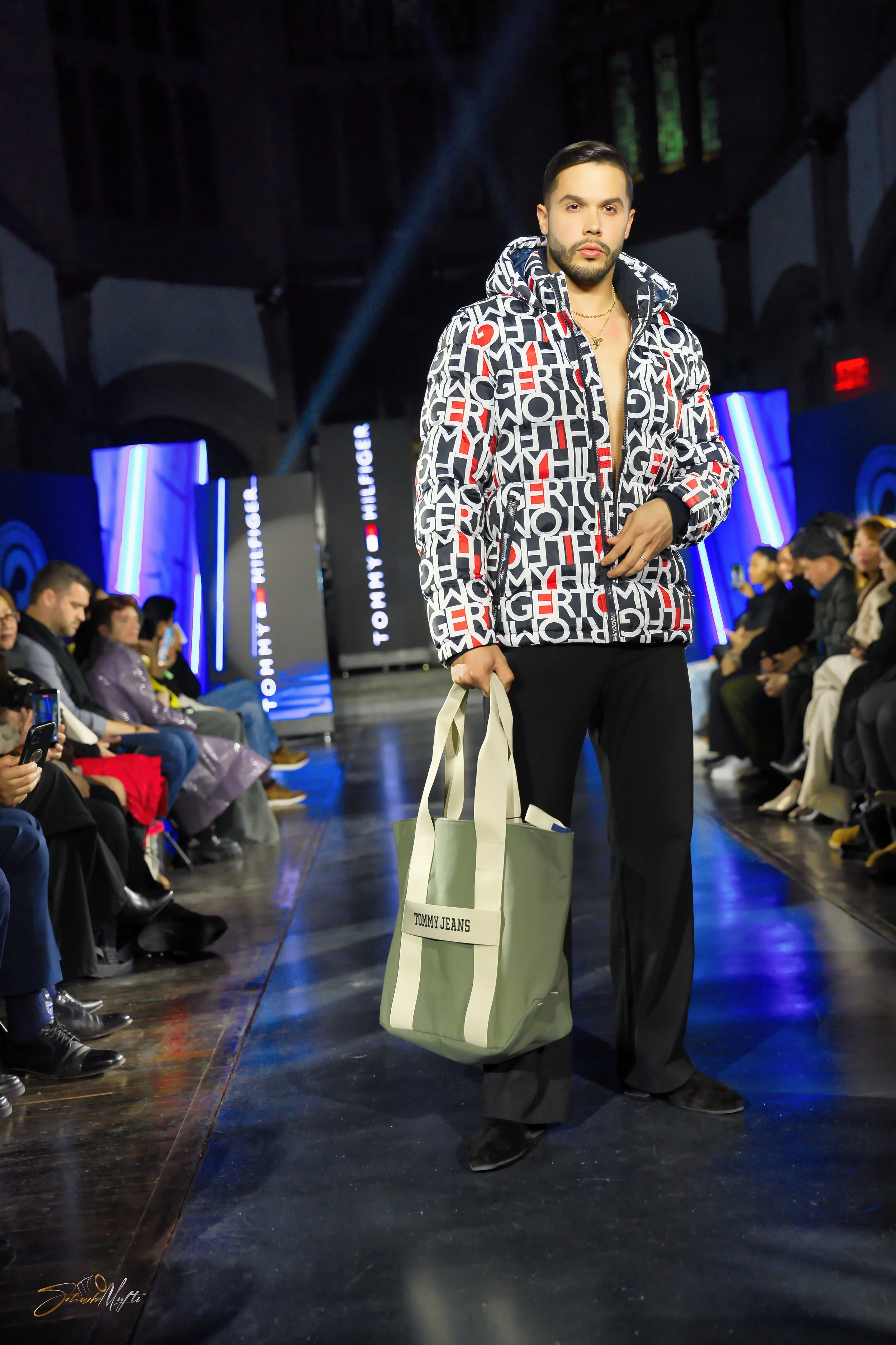 A male model walking on a runway at a Tommy Hilfiger fashion show, holding a Tommy Jeans tote bag, wearing a Tommy Hilfiger jacket with a bold logo pattern, with audience members seated on both sides.