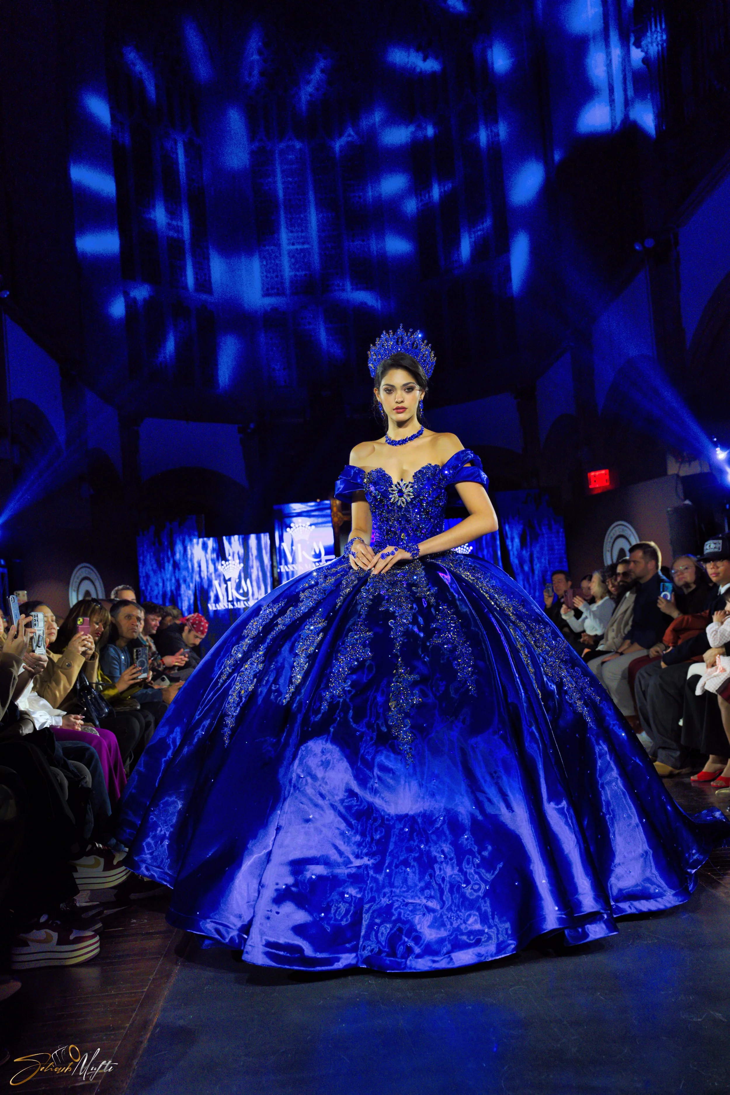 A woman walks down a runway wearing a blue ball gown and crown, surrounded by an audience taking pictures, with blue lighting and a digital backdrop.