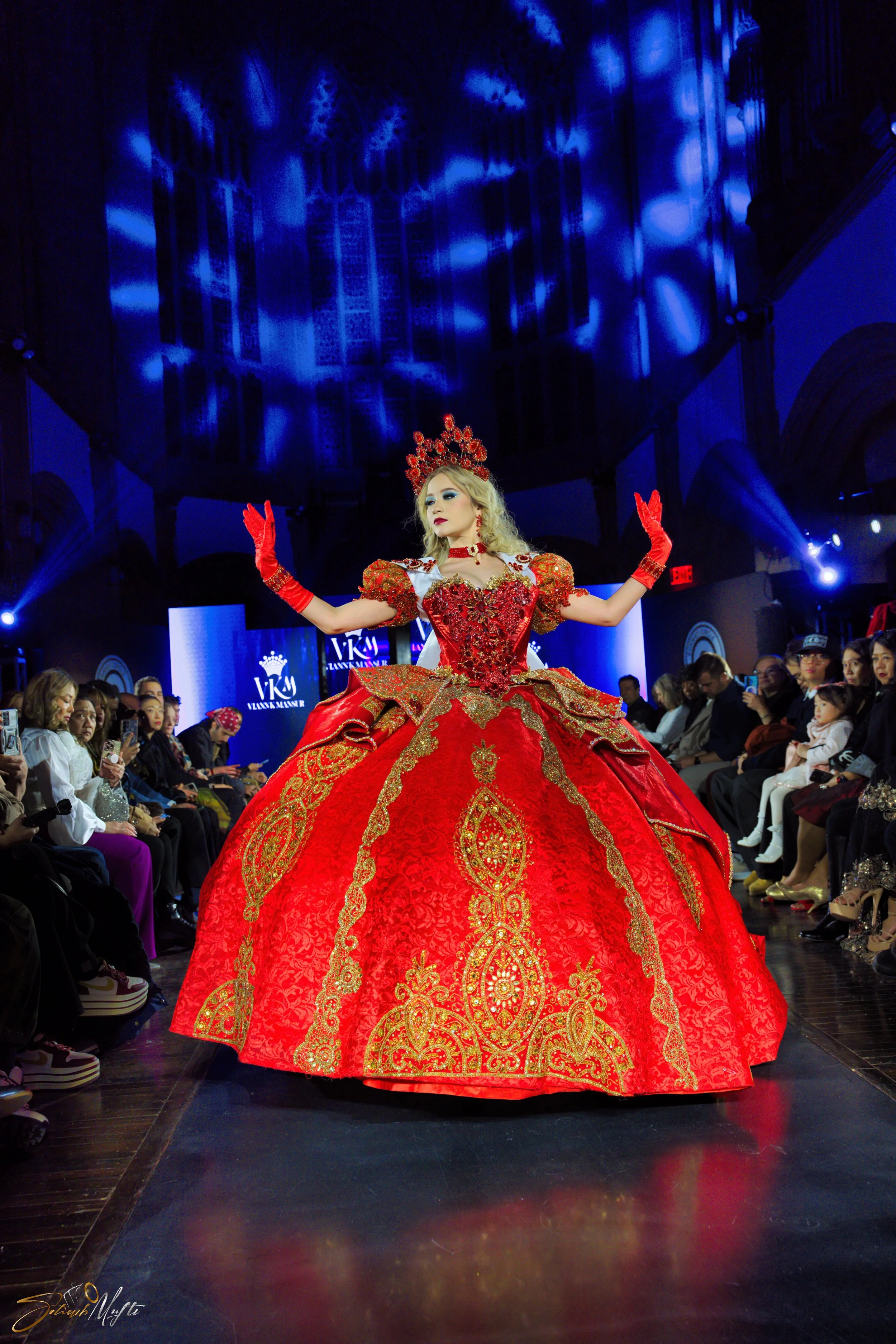 A female model walking on a runway during a fashion show, wearing a red and gold elaborate gown with puffed sleeves, gloves, and a matching headpiece, with audience members watching and taking photos.