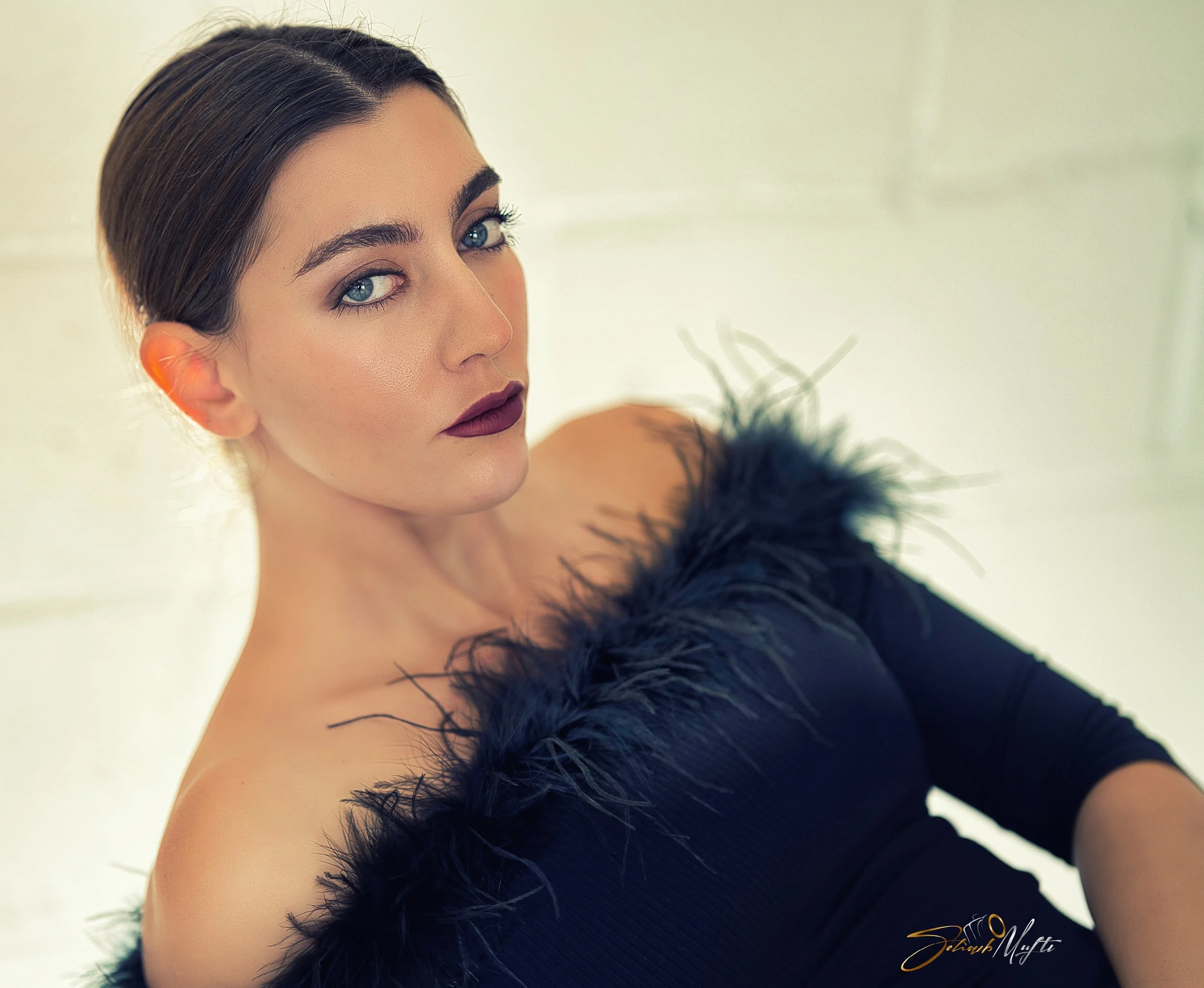 A woman with dark hair and blue eyes wearing makeup and a black dress with feathered shoulders, looking at the camera against a light background.