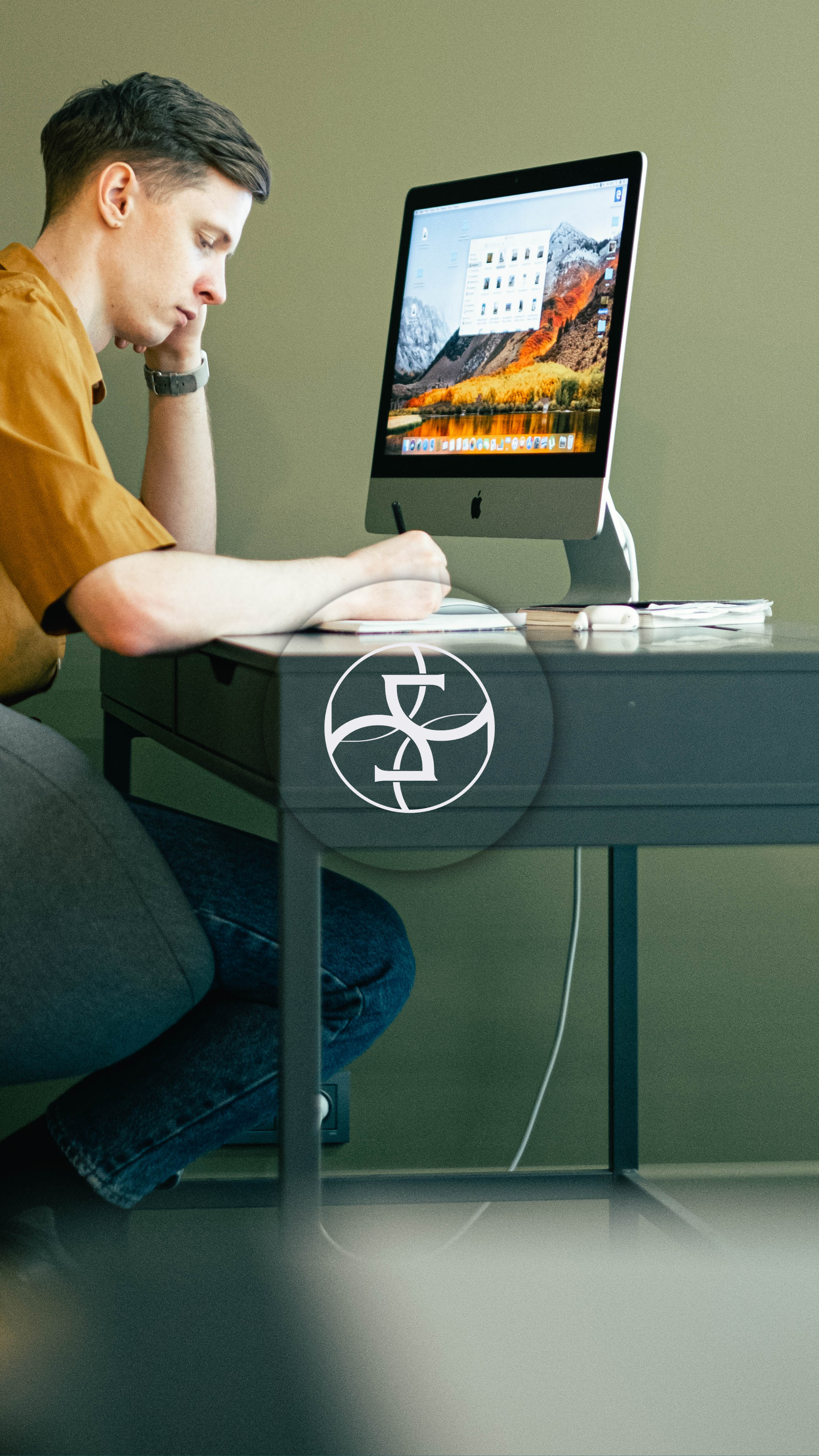 Young man with brown hair, wearing a brown shirt, sitting at a desk and working on a notebook while looking at a computer monitor.