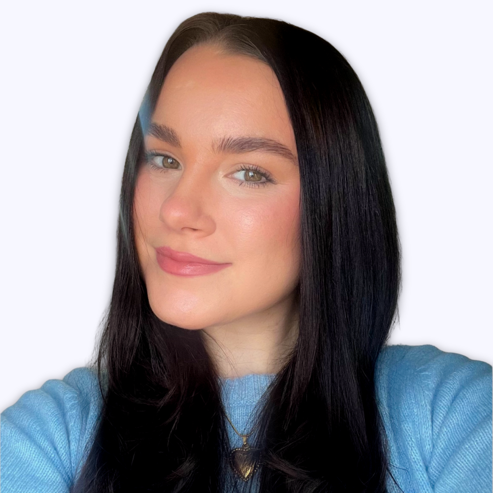A young woman with long dark hair, hazel eyes smiling softly, wearing a light blue top and a gold heart necklace against a plain white background.