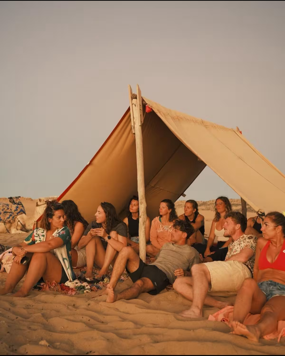 Group of people sitting and relaxing under a large canvas shade on sandy terrain during sunset.