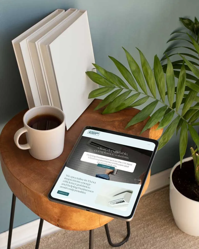 A wooden side table with a white coffee mug filled with coffee, four white books or binders leaning against a blue-green wall, a tablet displaying a website, a potted plant with green leaves, and beige carpet.