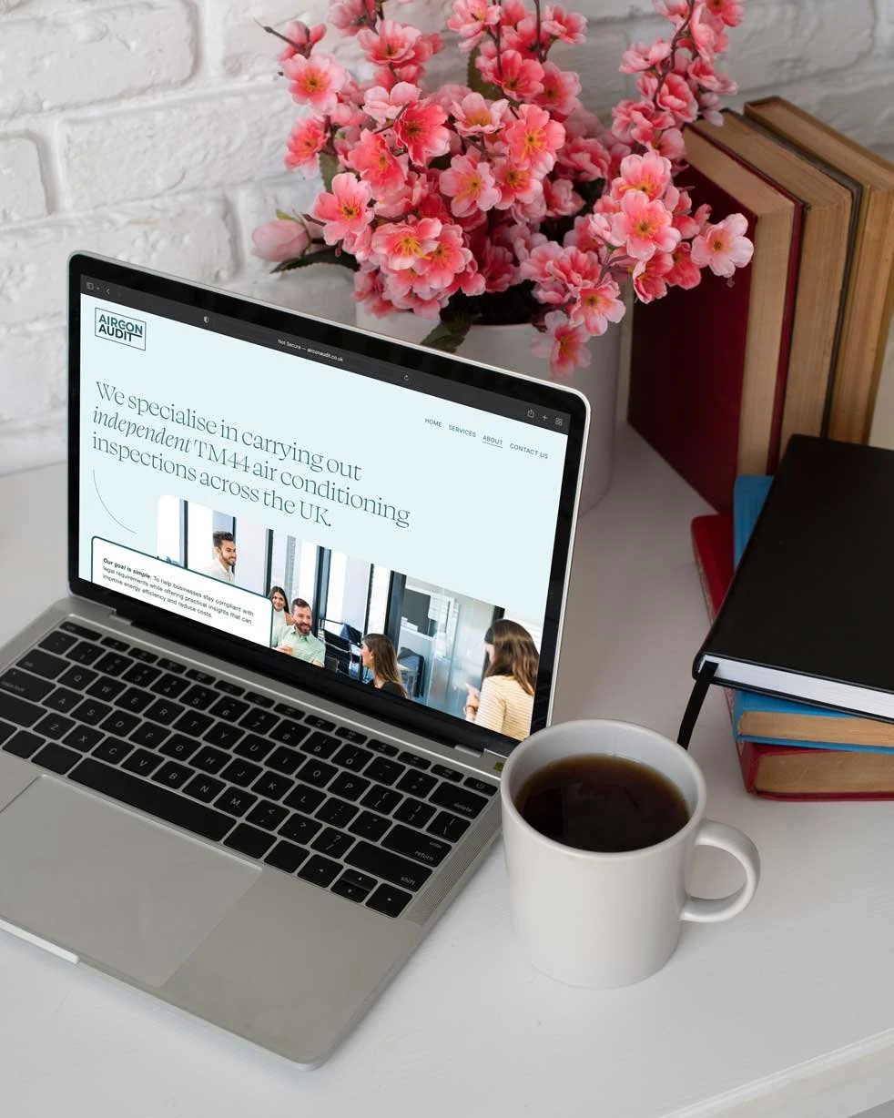 Laptop on a white desk displaying a website about air conditioning inspections in the UK, with a white coffee mug, a stack of books, and pink flowers in the background.