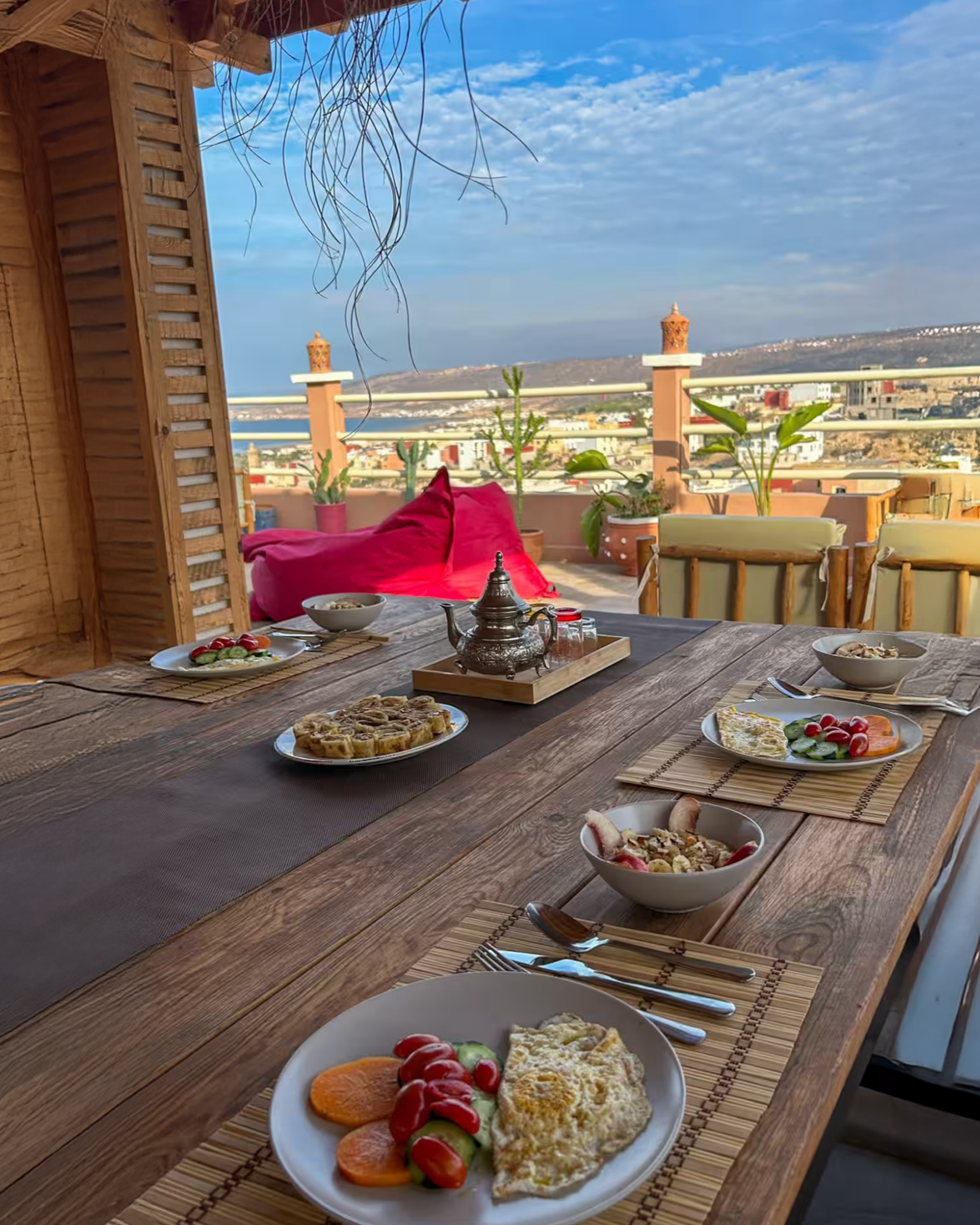 A rustic wooden dining table set with plates of breakfast, including eggs, vegetables, and bread, on a terrace with potted plants and a scenic view of a city and hills in the background.