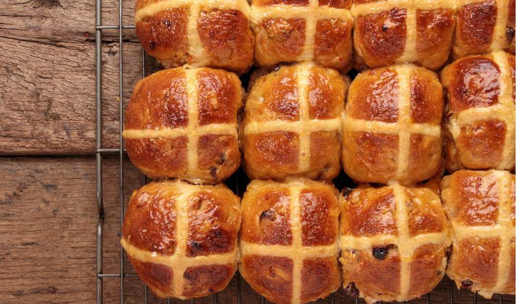 Close-up of freshly baked hot cross buns with golden-brown tops and white cross markings on a wire cooling rack on a wooden surface.