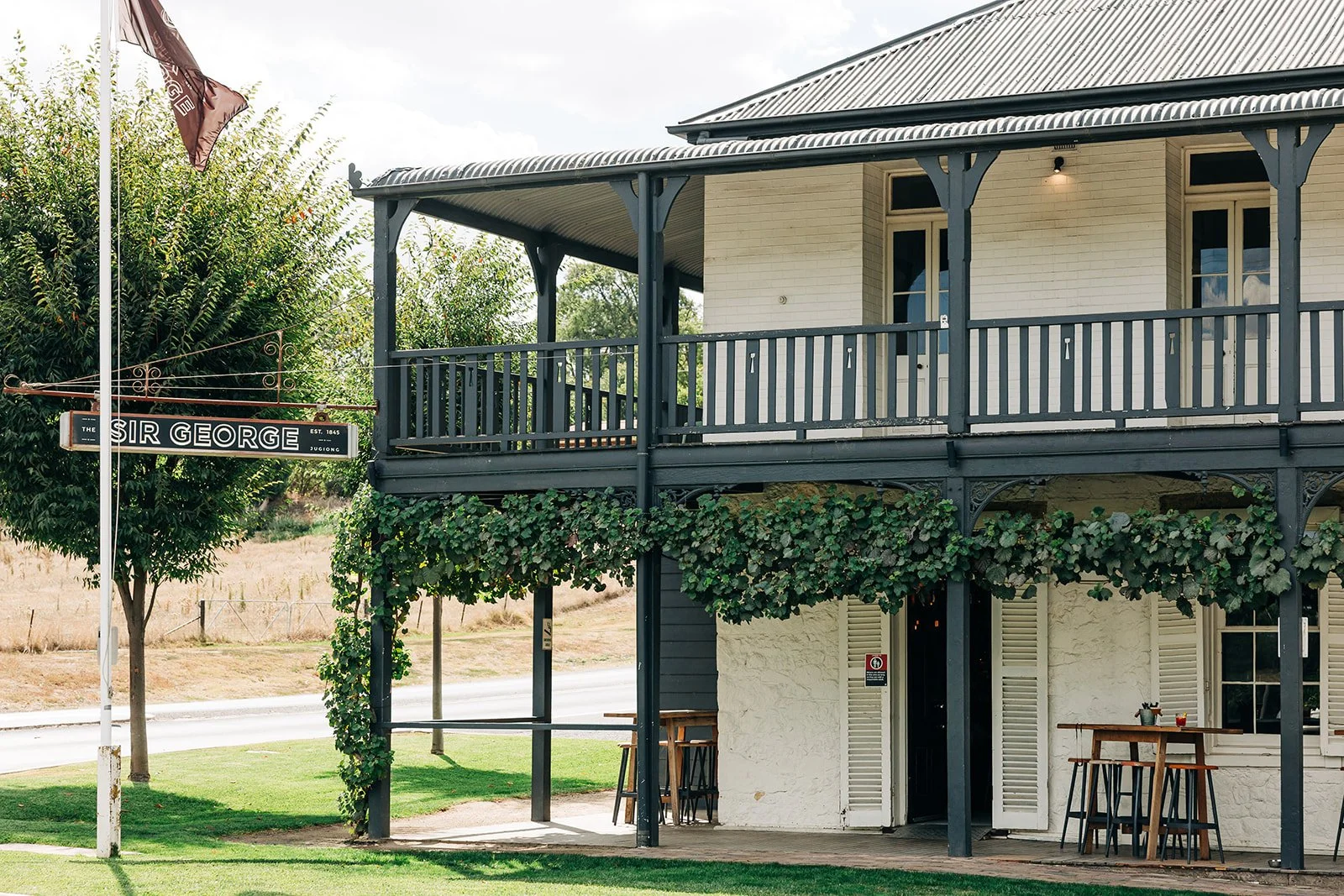 A two-story building with a balcony on the upper level, painted white with dark trim, surrounded by greenery. There is a sign that reads "The Sir George" on a flagpole near the building.