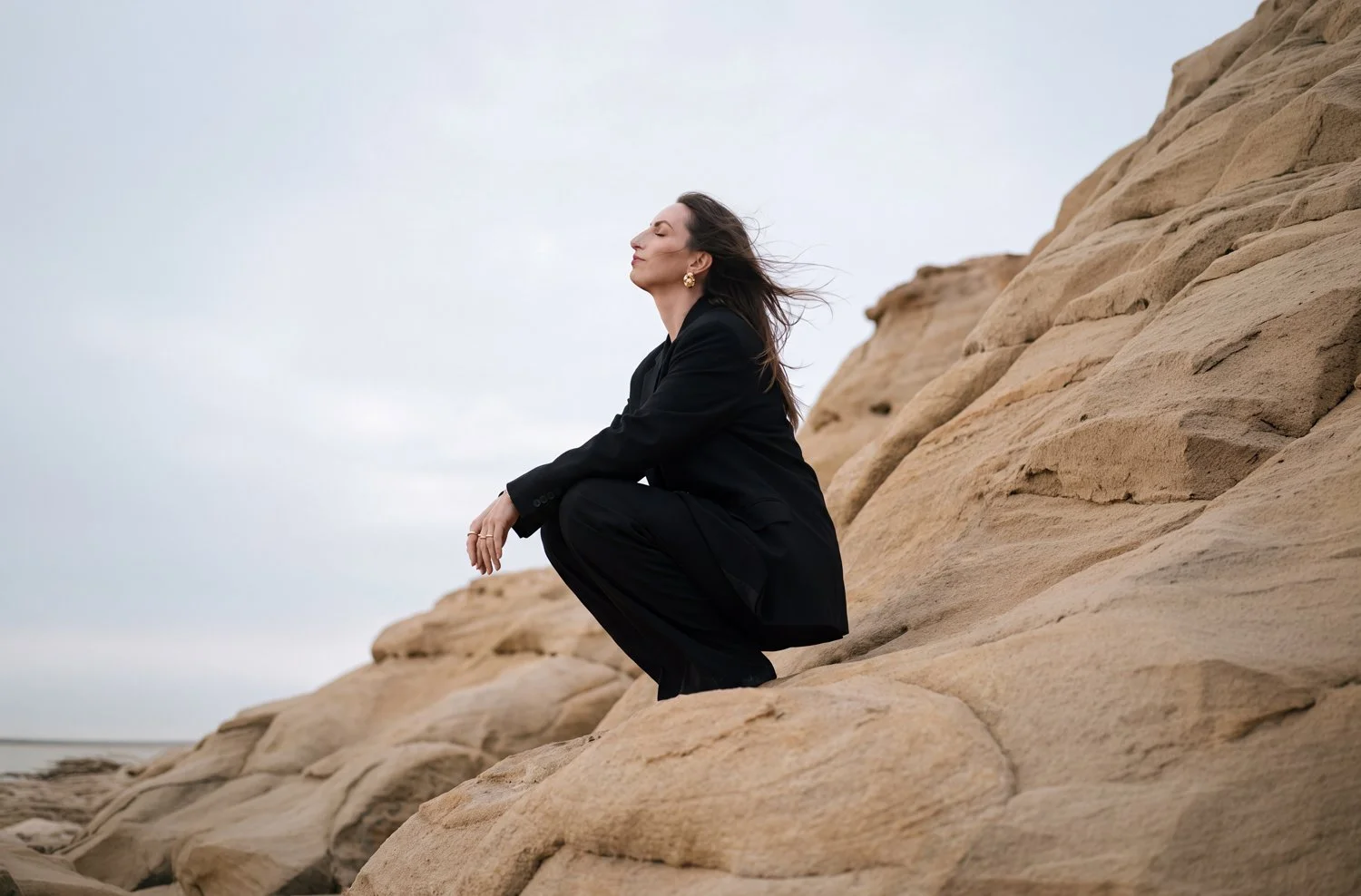 Carola Jansen im schwarzen Anzug hockt mit geschlossenen Augen auf hellen Sandsteinklippen am Meer, der Wind weht durch ihre Haare