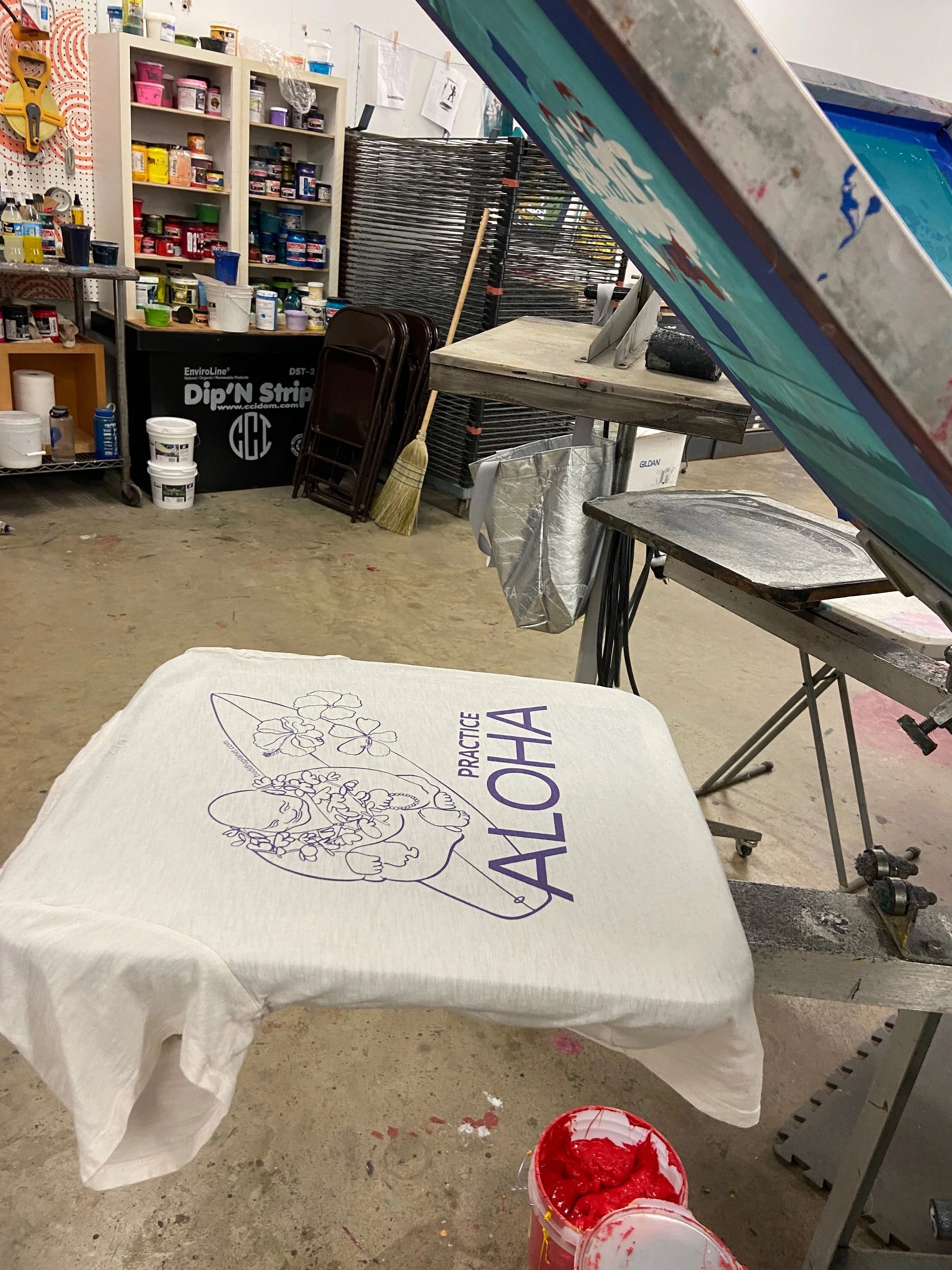 An art studio workspace with a fabric blanket draped over a table that has 'ALOHA' and a floral design printed on it. Worktables, shelves with paint containers, a broom, and art supplies are visible in the background. There are open paint buckets in the foreground.