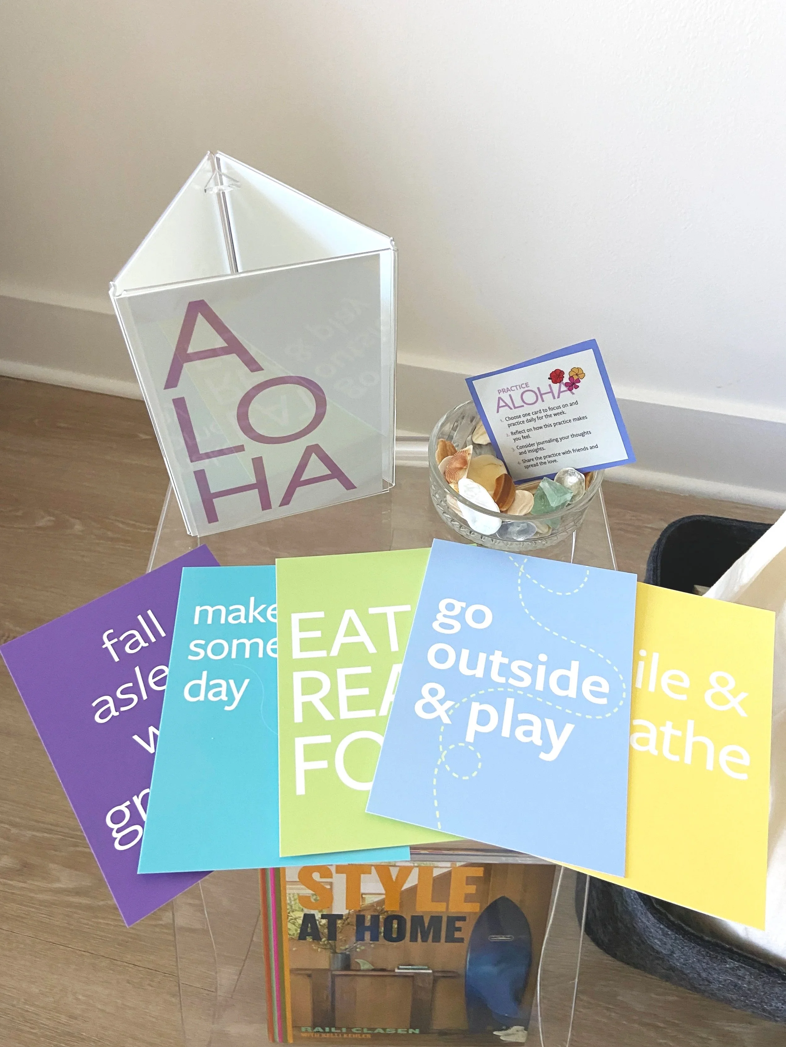 Colorful signs with outdoor and activity prompts such as 'fall', 'make some', 'EAT READ', 'go outside & play', and 'smile & breathe' arranged on a transparent table, with a bowl of seashells and a card about practicing aloha nearby.